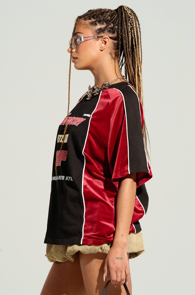 A woman with braids and wearing reflective sunglasses is seen in a side profile. She is dressed in THE AMERICAN JERSEY, which features colorblock paneling in black and red, complemented by beige shorts and a chunky necklace. She holds a small accessory in her hand and stands against a plain background.