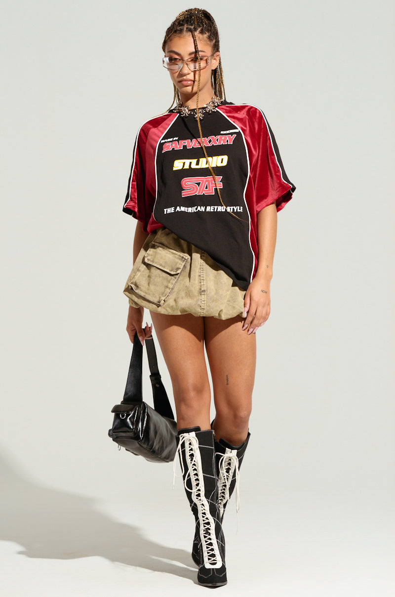 A person stands facing forward, wearing braided hair, glasses, THE AMERICAN JERSEY in red and black with colorblock paneling, a beige mini skirt, and black knee-high boots with white laces. They are holding a black handbag in their left hand.