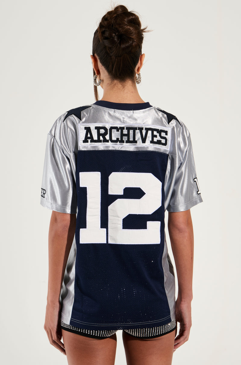 A person stands with their back to the camera, showcasing a sporty chic **SLICK JERSEY** that displays the text "ARCHIVES" on the upper back and the number "12" beneath it. This stylish jersey boasts a colorblock pattern in dark blue and silver. The individual has their hair up and is also wearing shorts.