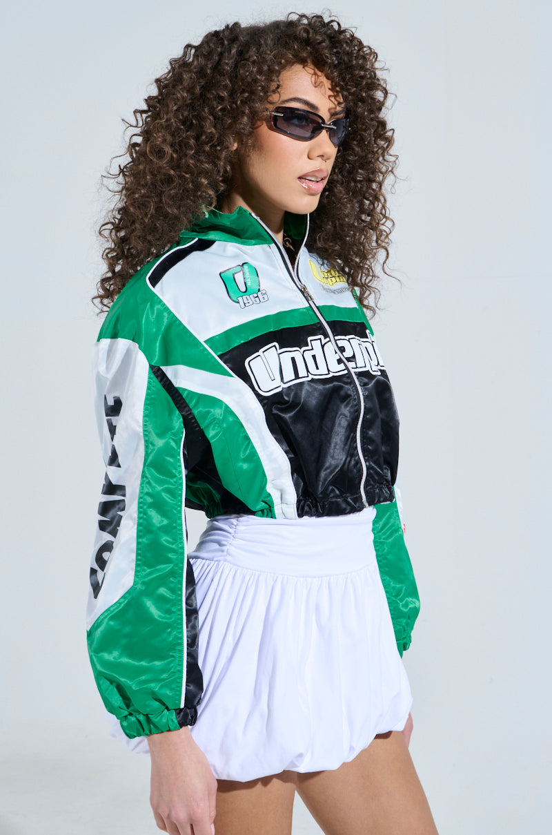 A woman with curly hair models the GO TIME EMBROIDERED BOMBER in green, black, and white with racing graphics. She pairs it with white high-waisted shorts and dark sunglasses, creating a sporty look against a plain light background.