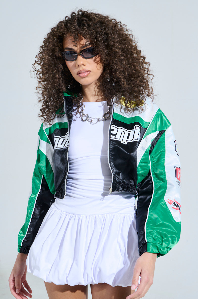 A woman with curly hair and sunglasses wears the GO TIME EMBROIDERED BOMBER, featuring racing-inspired graphics in green and black. Her outfit is completed with a white top and matching skirt as she stands against a plain background, gazing forward.