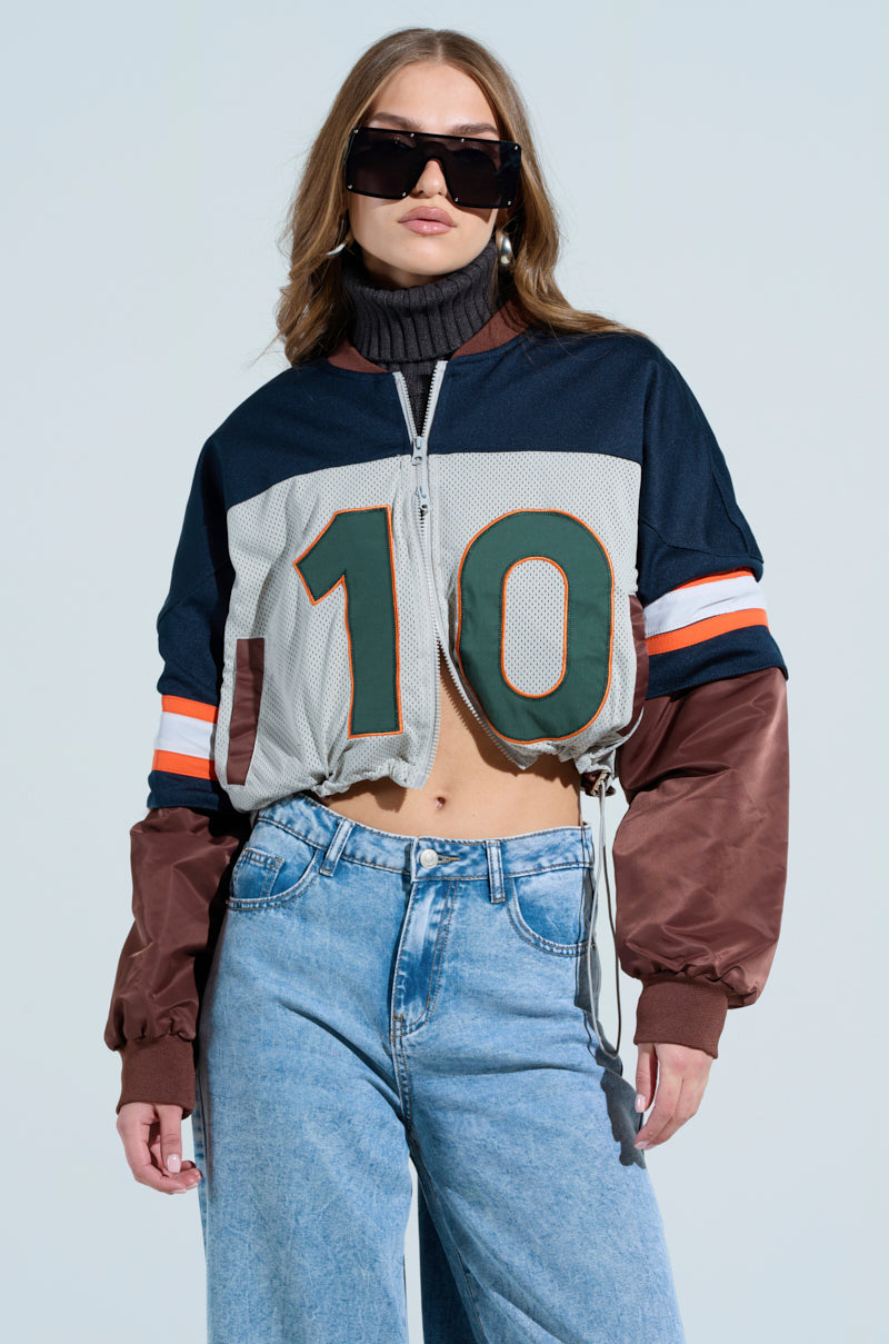 Wearing oversized sunglasses and the TOP TEN BOMBER—a varsity-style cropped mesh sports jersey with number 10 and blue, brown, and orange stripes—a woman poses confidently. It’s paired with loose jeans, her long wavy hair flowing over a grey turtleneck underneath.