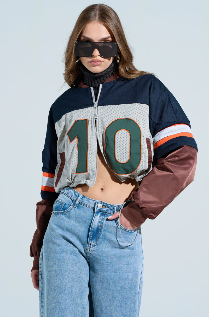 Wearing the Top Ten Bomber, a cropped, varsity-inspired jersey with the number "10" in black, white, green, orange, and brown. Paired with oversized sunglasses and light blue jeans against a light gray backdrop.