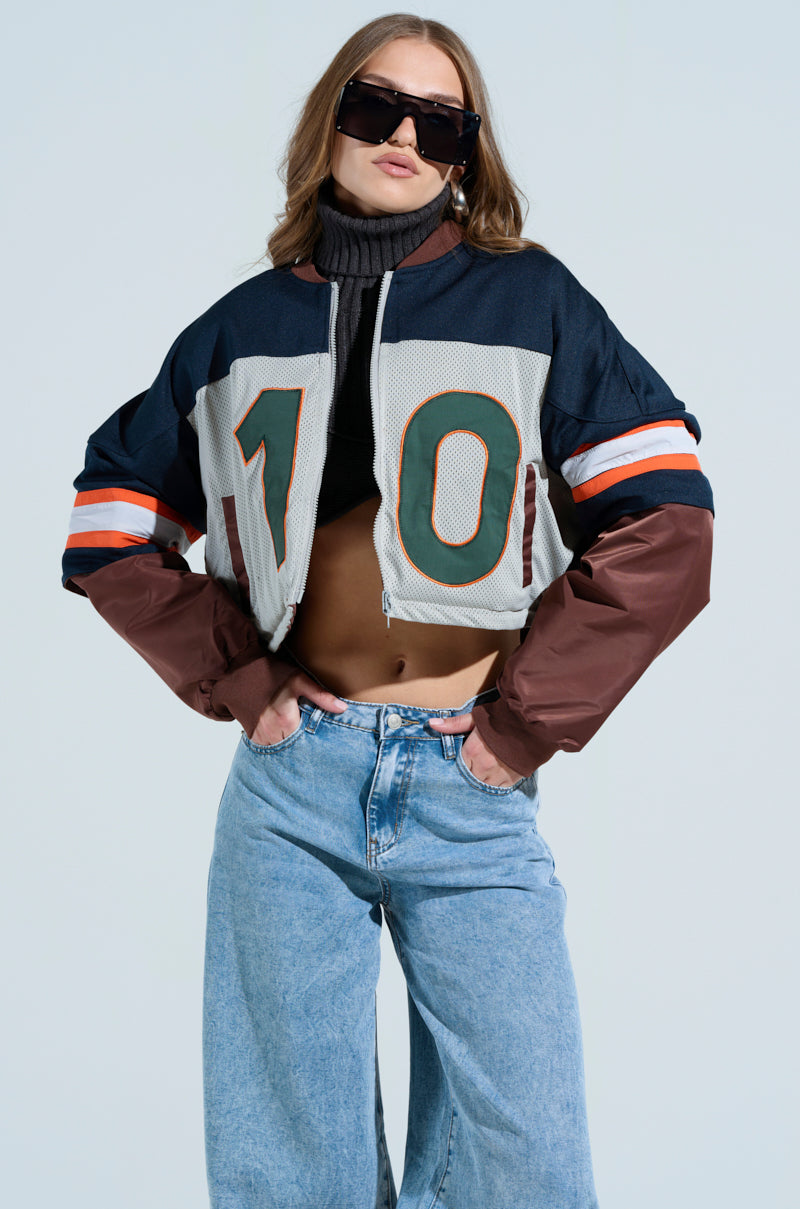 A person confidently poses in dark sunglasses, a high-neck black crop top, and the TOP TEN BOMBER jacket with "10." Their baggy light blue jeans enhance their long hair's cool vibe as they step forward effortlessly.
