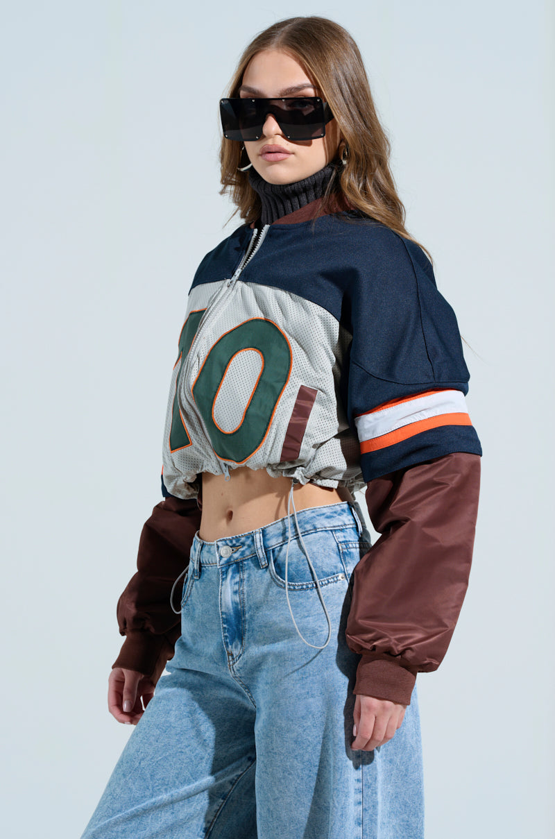 Wearing oversized sunglasses, a woman showcases the Top Ten Bomber style with a varsity-inspired, cropped multicolor jacket featuring "10." The sporty ensemble presents navy, brown, and gray tones with white and orange stripes, complementing her light blue jeans against a plain backdrop.