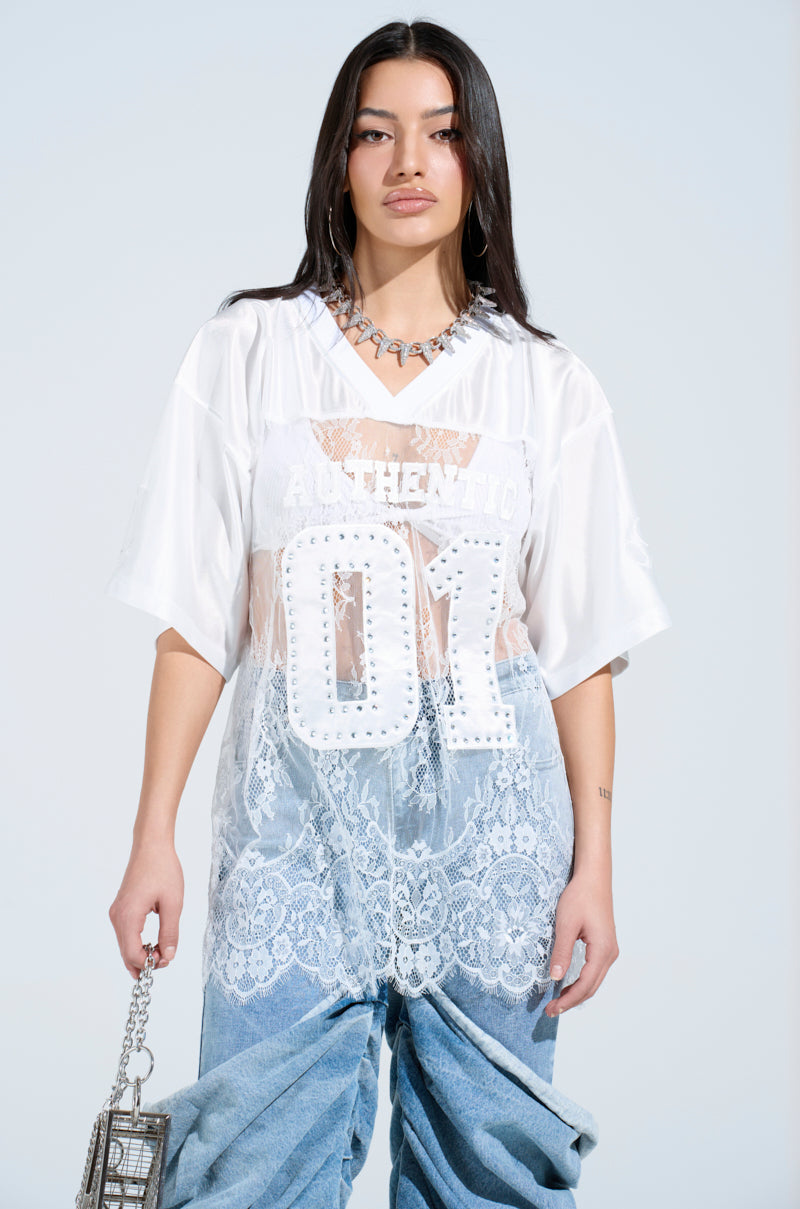 A woman in a sheer white "Authentic 01" jersey with floral lace details and oversized fit pairs it with baggy jeans. She holds a small metallic chain bag, her long hair flowing down with a silver necklace set against the light blue background.
