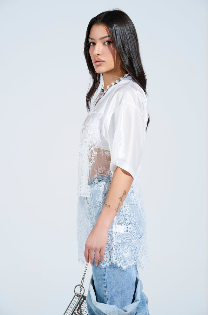 A person with long dark hair stands sideways, wearing a charming lace jersey dress over jeans, holding a rhinestone-embellished silver bag. Tattoos adorn their arms against a plain white background.