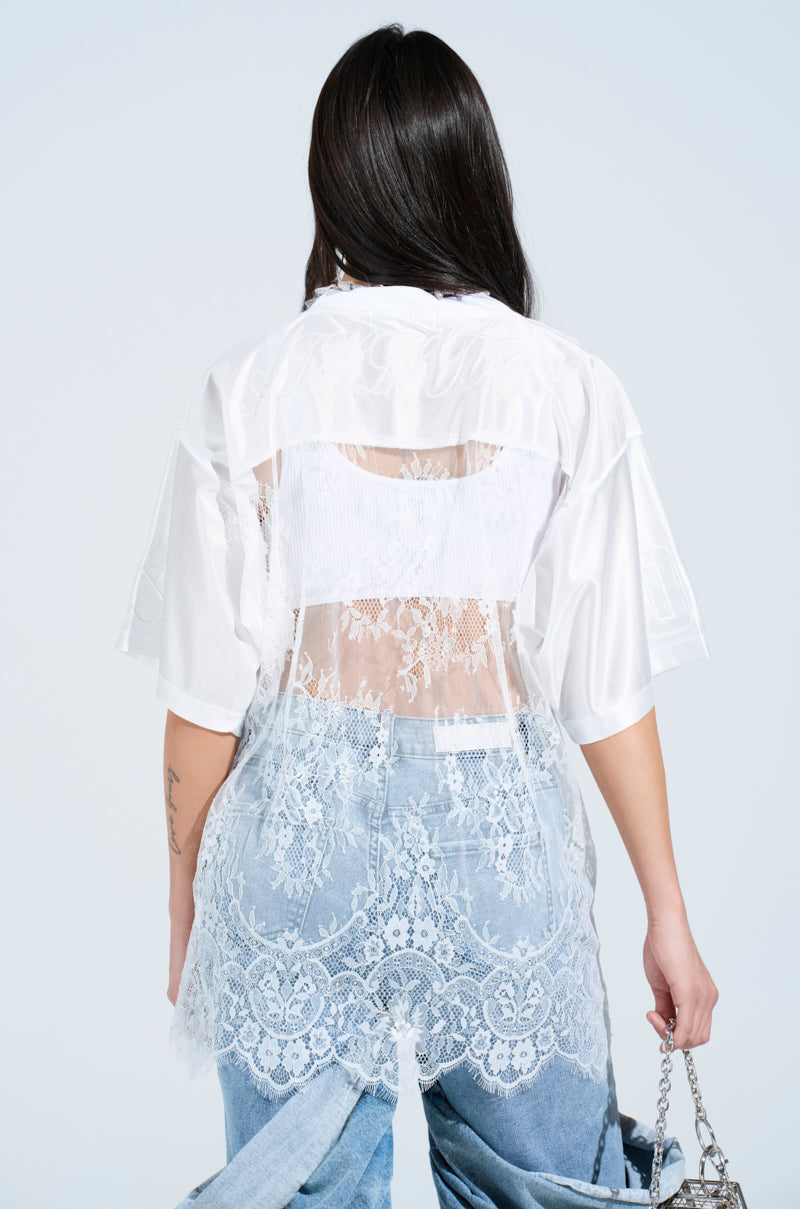 A person with long dark hair is wearing the CHARMING LACE JERSEY DRESS, featuring floral lace patterns over a white shirt, paired with ripped blue jeans. They hold a silver chain accessory, showcasing intricate designs at the back.