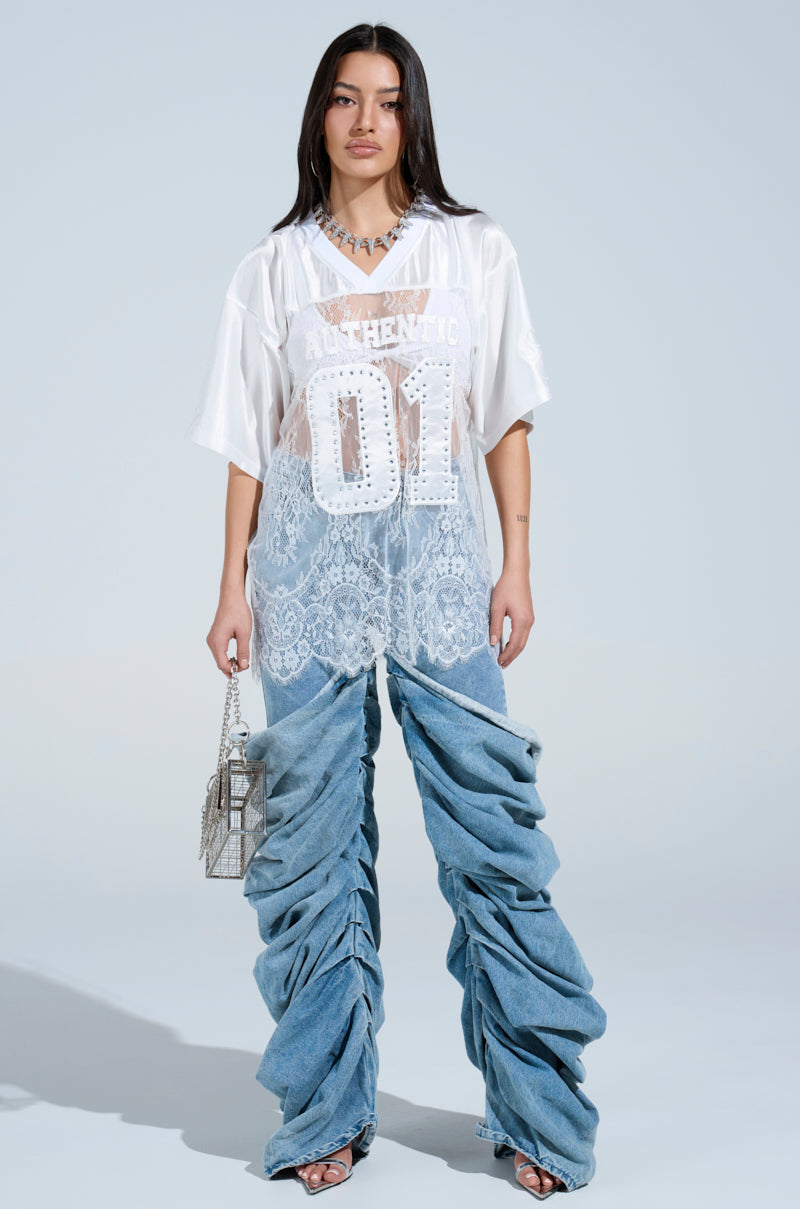 A confident woman stands against a light backdrop, wearing an oversized sheer white top with "AUTHENTIC 01" and ruched denim jeans. She carries a small silver handbag and dons open-toe shoes, giving off a chic vibe similar to the CHARMING LACE JERSEY DRESS.