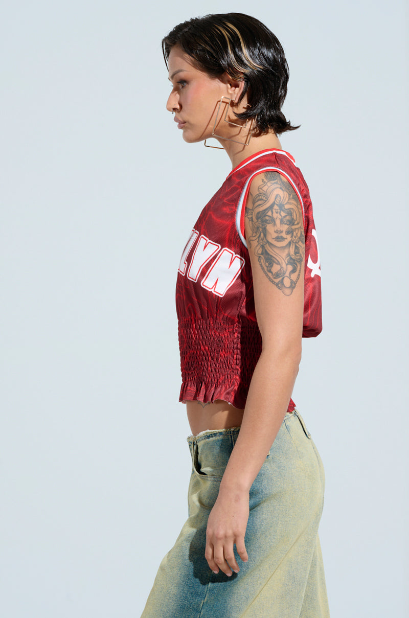 A person with short, dark hair and a side profile is wearing the "Youre A Rookie Jersey in Red" featuring white Brooklyn graphic lettering and a long denim skirt. They have a large tattoo on their upper arm and face away from the camera against a plain background.