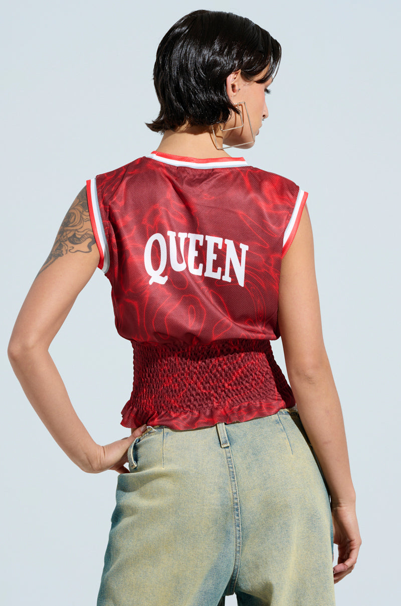 A person with short dark hair and a tattoo stands with their back to the camera, wearing the sleeveless YOURE A ROOKIE JERSEY IN RED featuring "QUEEN." Paired with light denim jeans against a plain background, it creates a strikingly balanced look.