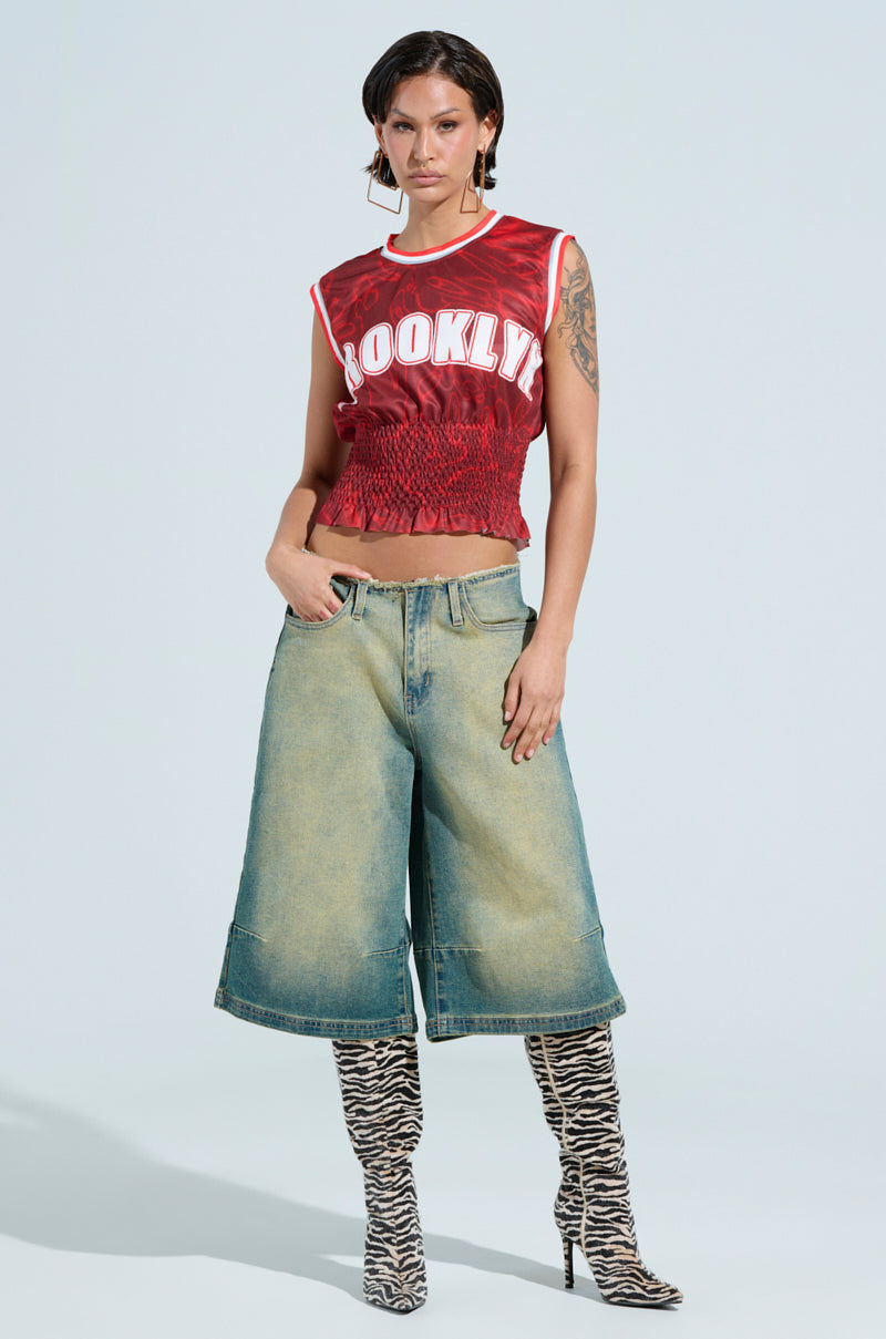 Against a light blue background, someone showcases the YOURE A ROOKIE JERSEY IN RED with "Brooklyn" displayed prominently, teamed with wide-leg denim shorts and zebra-patterned knee-high boots. Their short hair and arm tattoos enhance the edgy style.