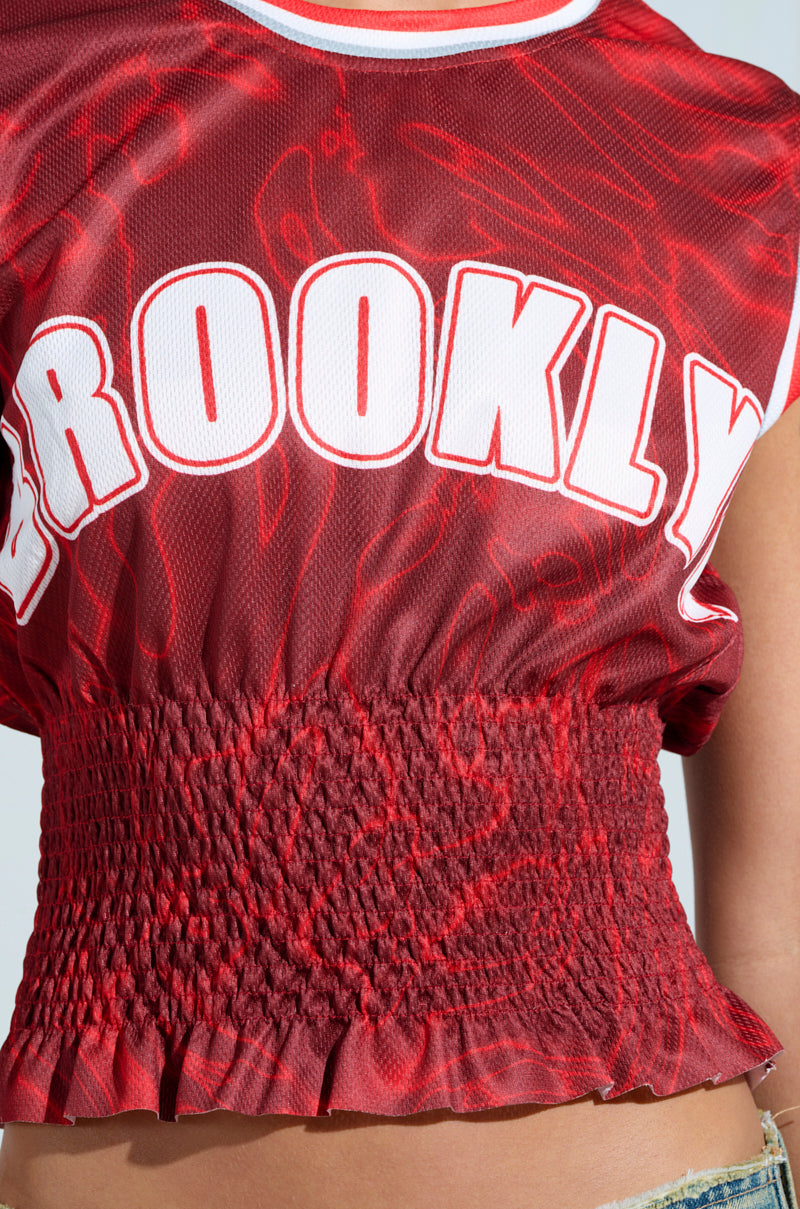 Close-up of a person wearing the YOURE A ROOKIE JERSEY IN RED, featuring a Brooklyn graphic in large white letters. The sleeveless crop top has wavy jersey fabric and a ruffled hem, with part of their arm and denim shorts visible.