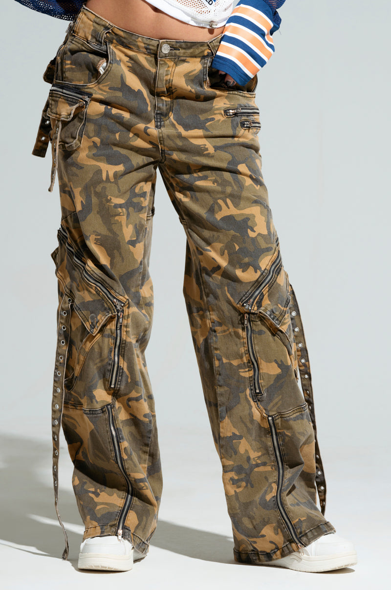 A person wearing the JUNGLE PANT, a high-rise wide-leg camouflage cargo with silver buttons and ties, paired with white sneakers. The pants provide a wide-leg fit and multiple pockets for both style and functionality.