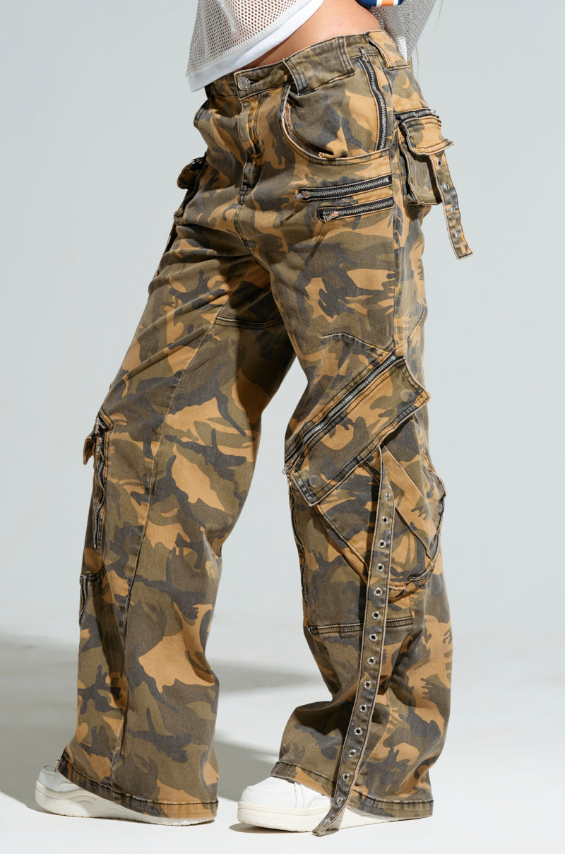 Someone sporting the JUNGLE PANT, a pair of high-rise wide-leg camouflage cargo pants adorned with multiple pockets and metal eyelet details, stands sideways against a plain, light-colored background. These loose-fitting pants exude a laid-back style.