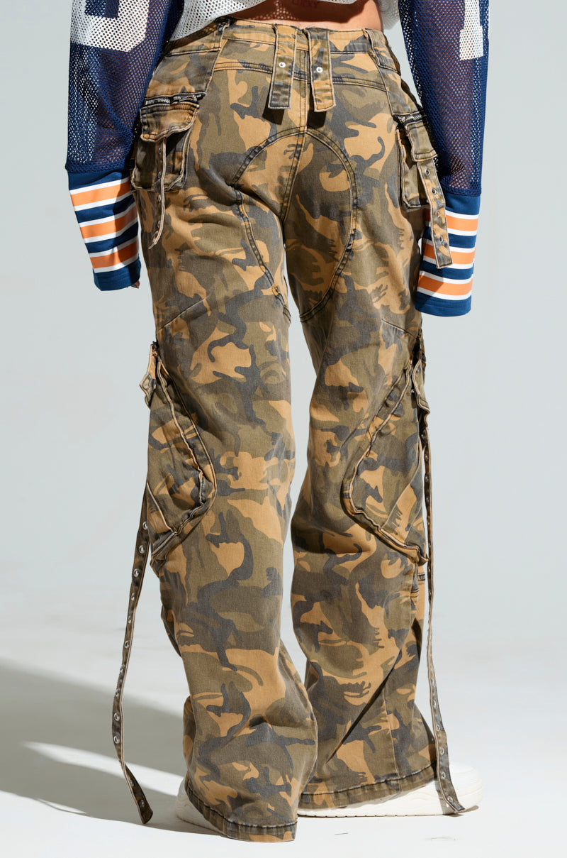 The JUNGLE PANT, a pair of high-rise wide-leg camouflage cargo pants with strap details, paired with a blue mesh top and white sneakers, perfectly embodies a laid-back streetwear vibe. The image captures the lower half of the body from the back, highlighting the outfit's urban flair.