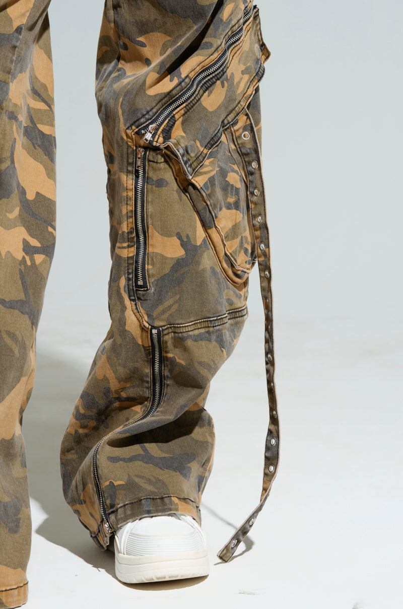 A close-up shows a person wearing the JUNGLE PANT, high-rise wide-leg camouflage pants featuring zippers, pockets, and extended straps, paired with white sneakers. The rugged design stands out against a plain gray background.