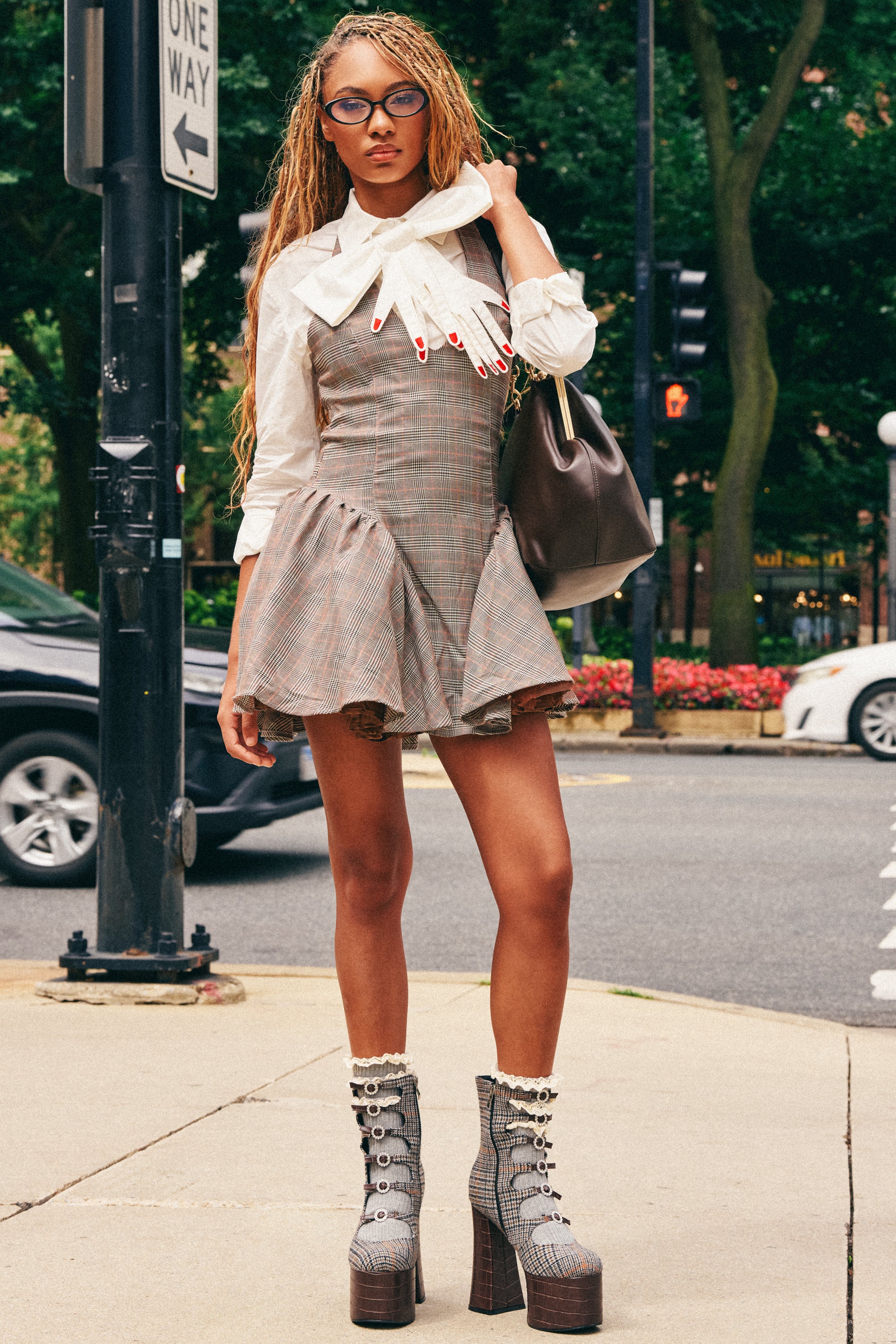 A person stands on a city sidewalk wearing glasses, the STYLE AND GRACE MINI DRESS IN PLAID layered over a white blouse, platform boots, and carrying a brown bag. Trees, traffic signs, and cars can be seen in the background.