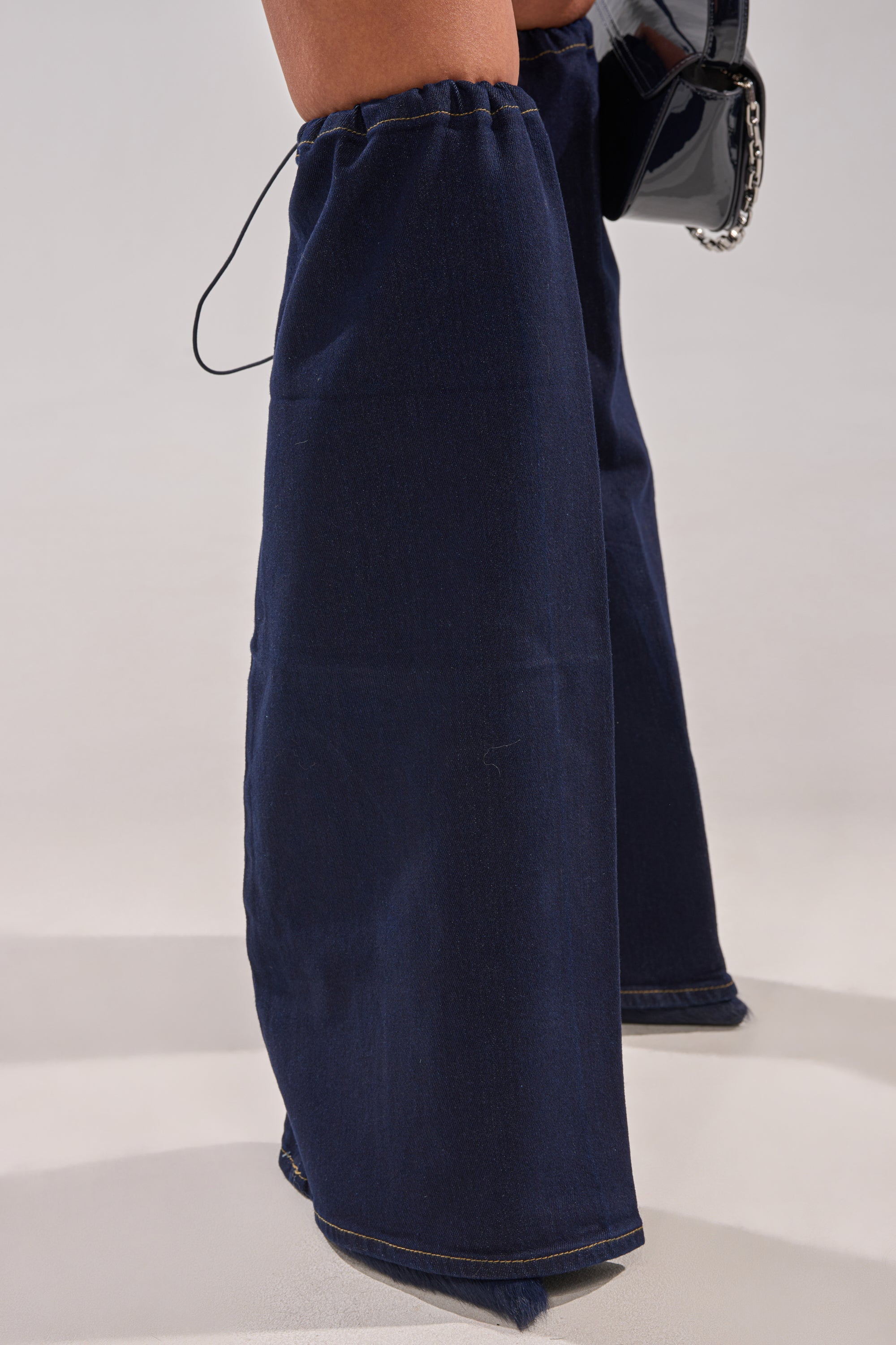 A person stands on a light surface wearing the BUSINESS MEETING CHIC BLAZER DRESS AND LEG WARMER SET IN DENIM, featuring ultra-wide dark blue denim pants that nearly cover the feet, paired with a shiny black handbag.