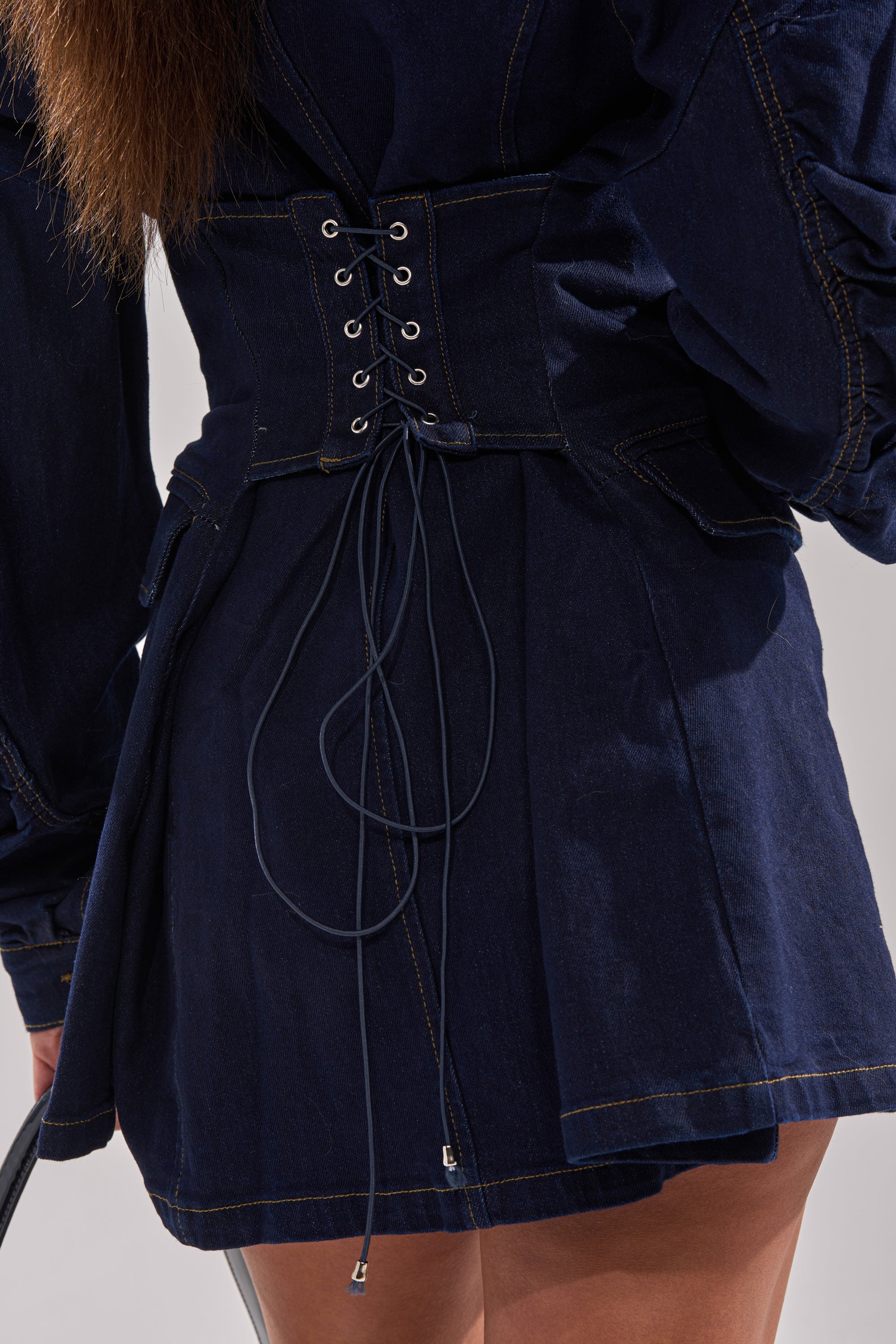 A close-up shows someone wearing the BUSINESS MEETING CHIC BLAZER DRESS AND LEG WARMER SET IN DENIM, featuring long sleeves, a lace-up corset back, prominent stitching, flared skirt, and holding a silver handbag.