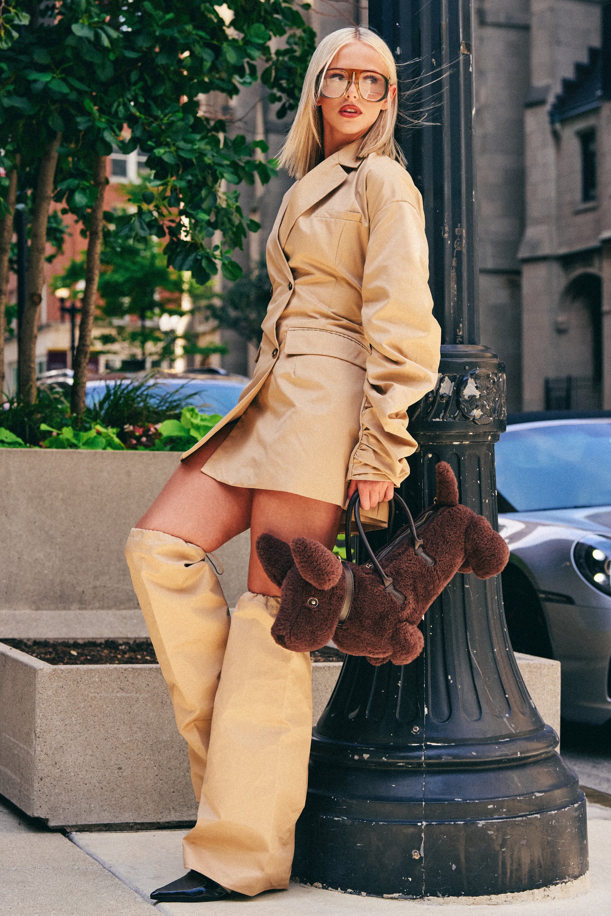 Wearing the BUSINESS MEETING CHIC BLAZER DRESS AND LEG WARMER SET IN TRENCH, a woman styled with oversized boots and clear glasses leans against a lamp post, carrying a brown dog-shaped plush bag—her look exemplifies modern workwear-inspired street style in an urban outdoor setting.