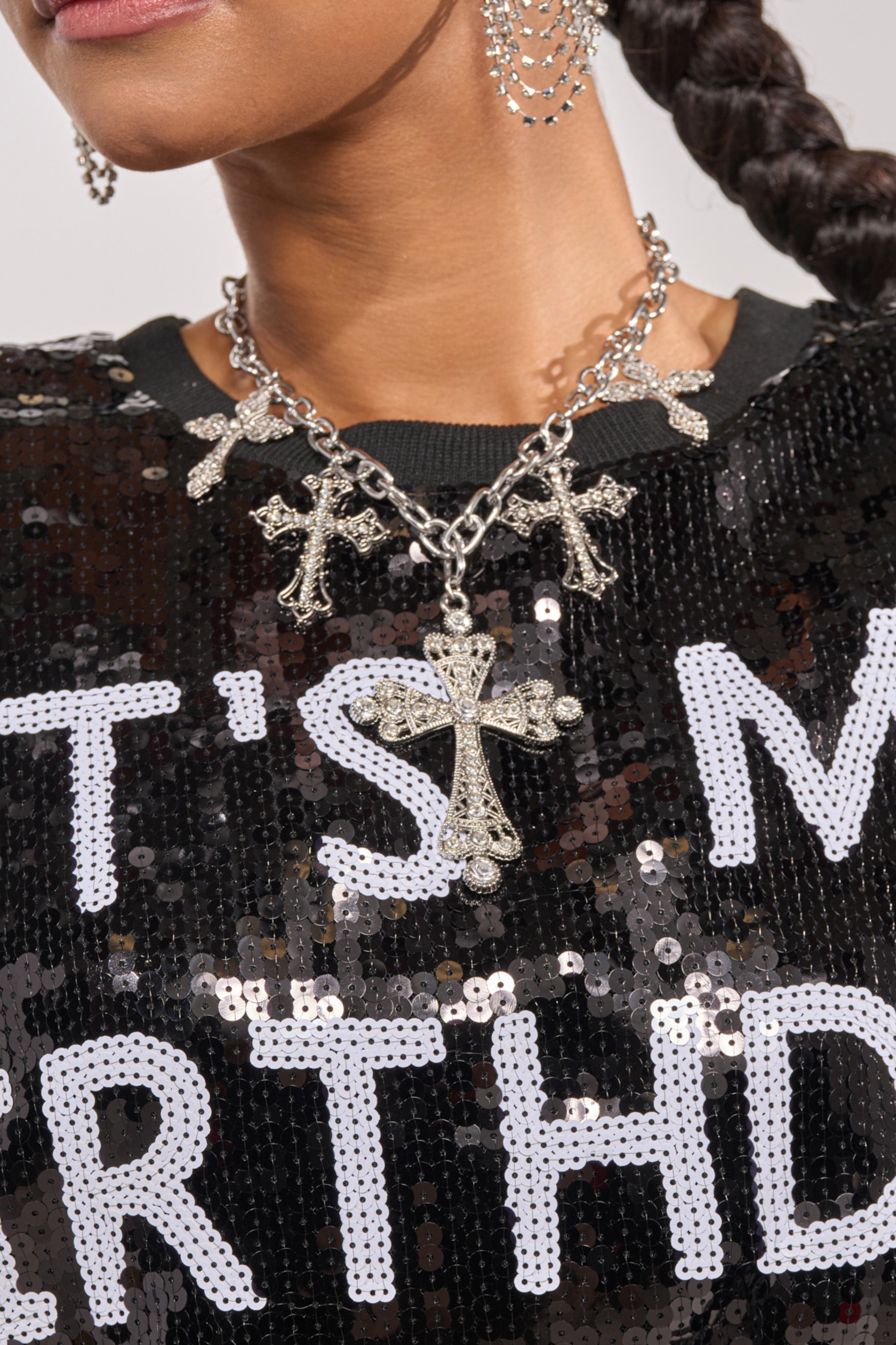 A woman wears a black sequin top with white block letters and the STAY BLESSED NECKLACE, a statement piece featuring multiple silver-tone cross charms, paired with matching cross earrings. 1