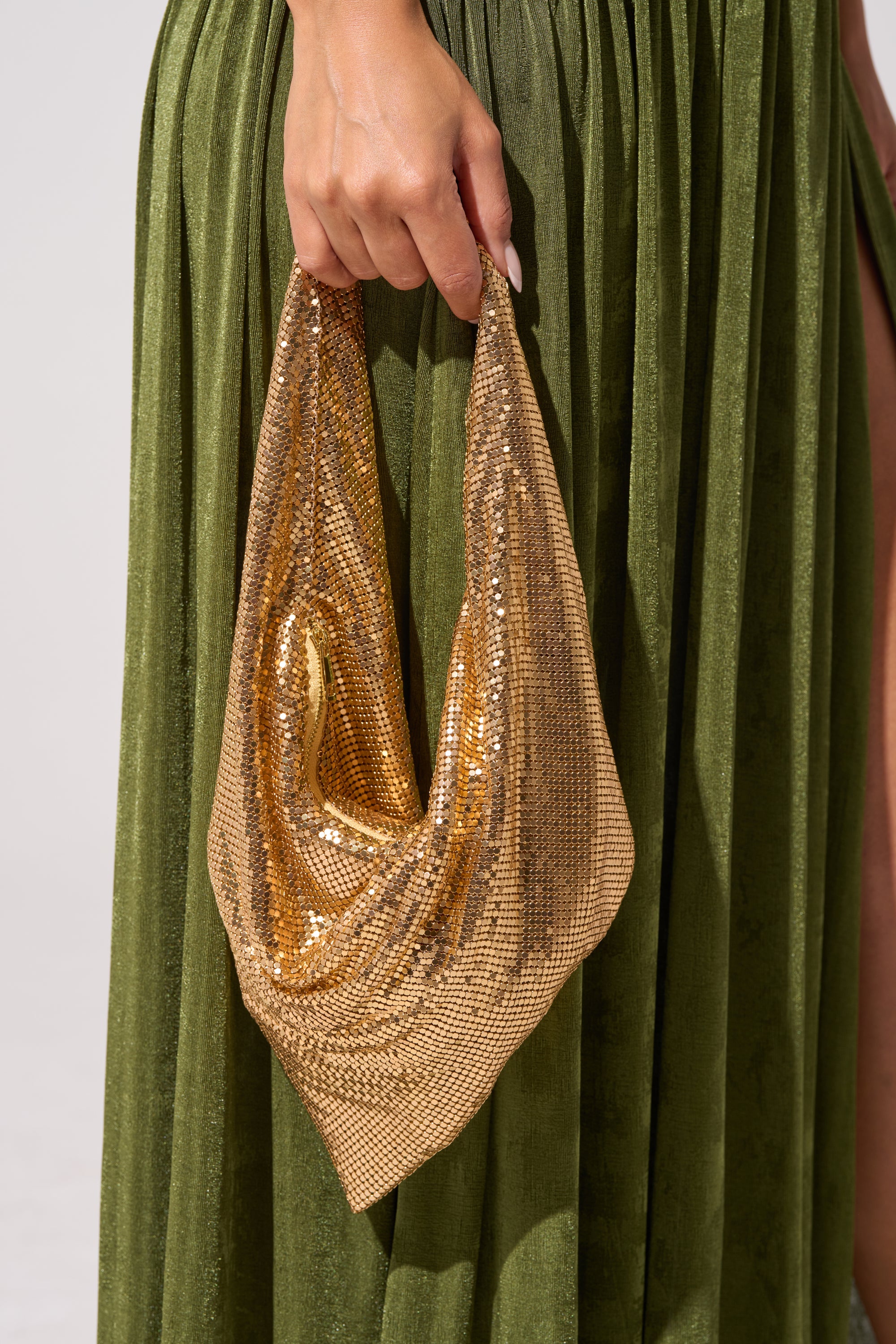A person in a green pleated skirt holds the MEET ME AT THE DISCOTHEQUE CHAINMAILL BAG, its sparkling rhinestone gold finish and draped design perfectly complementing their manicured nails. 0