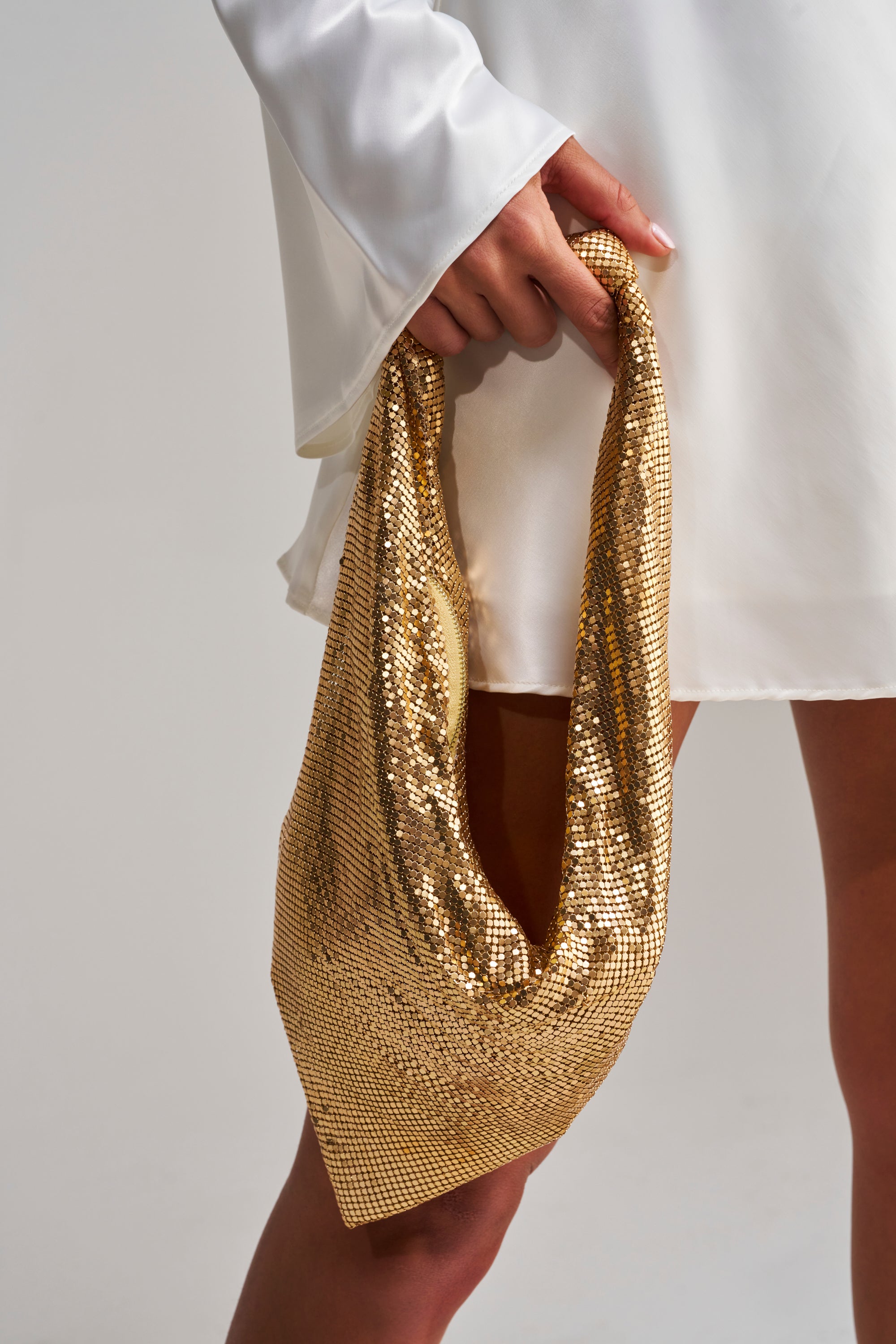 A person in a white dress holds the MEET ME AT THE DISCOTHEQUE CHAINMAILL BAG by their side against a plain background, adding a touch of sparkle to the simple scene.