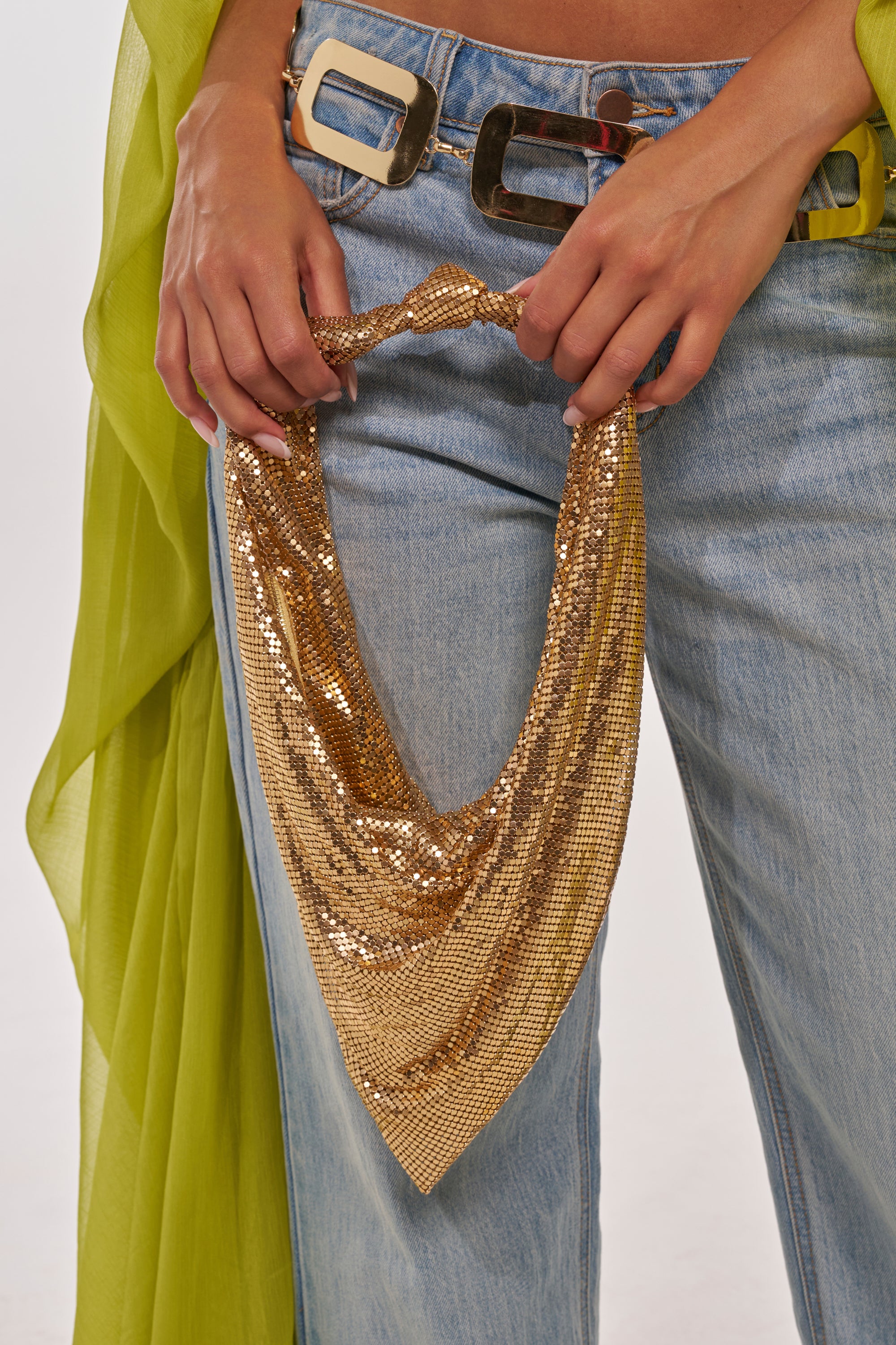 A person wearing light blue jeans, a sheer green top, and a wide belt holds the MEET ME AT THE DISCOTHEQUE CHAINMAIL BAG—a sparkling, triangular rhinestone gold handbag. 1