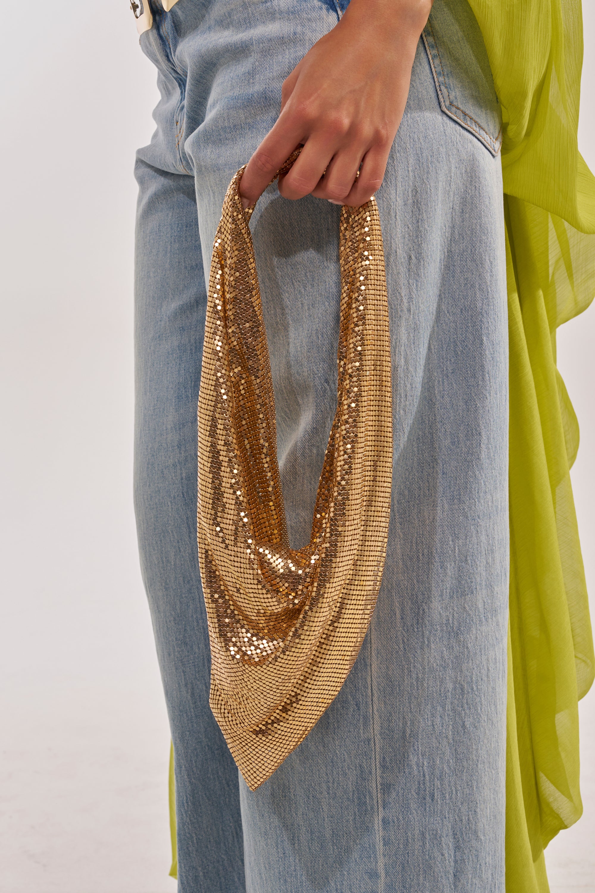 A person in light blue jeans and a green top holds the MEET ME AT THE DISCOTHEQUE CHAINMAILL BAG, a shiny gold rhinestone handbag, by their side against a plain white background. 2
