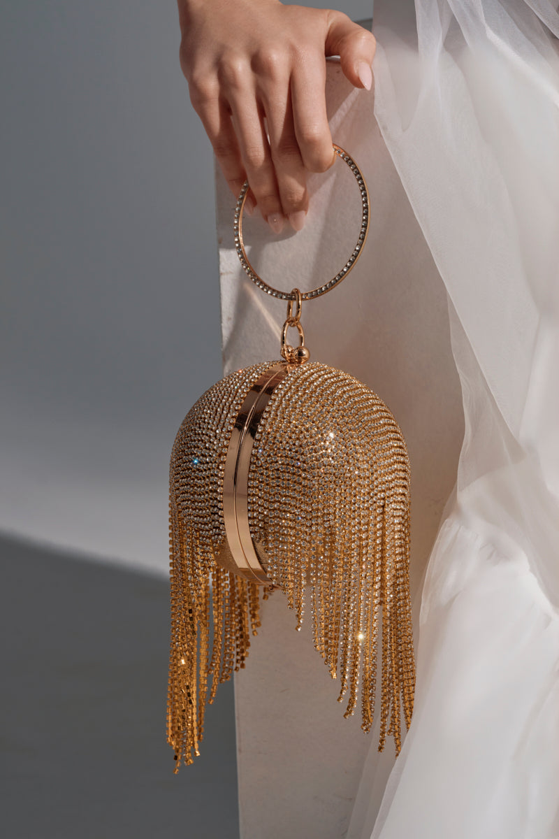 A hand holds the IM THE STAR BLING CLUTCH BAG, featuring a metallic gold base, long rhinestone chain link fringe, and a ring handle, set against white fabric in a neutral setting.