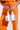 A person in an orange crop top and matching pants with drawstrings and bold white text on the leg holds the TO THE DISCO CHAINMAIL PURSE, a shimmering silver retro glam handbag with white handles, in front of their waist.