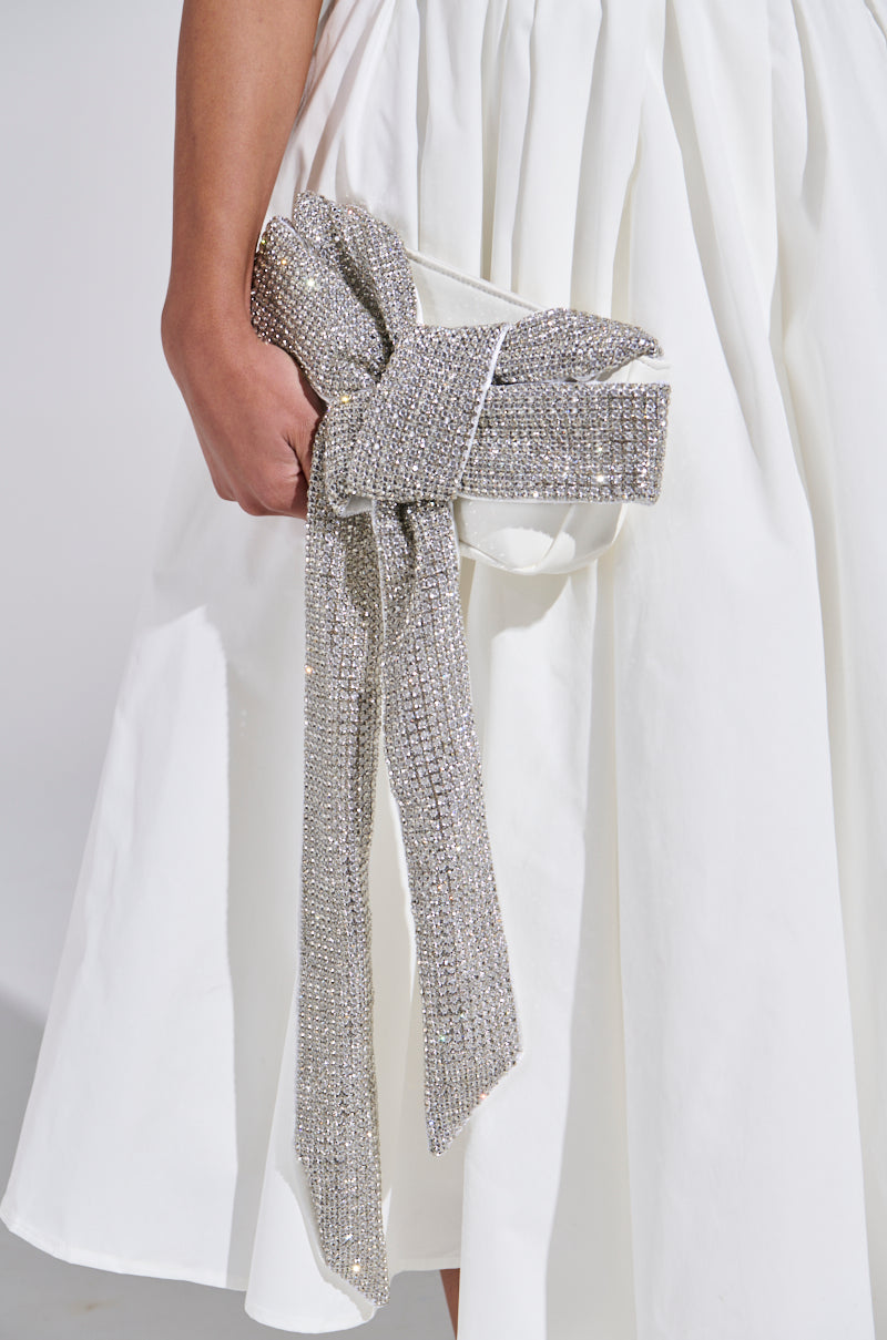 A person in a white dress holds the TIE IT UP RHINESTONE CLUTCH, a stunning faux leather accessory shaped like a sparkling silver bow. This clutch adds glamor to any evening look, while the neutral background highlights its elegance.