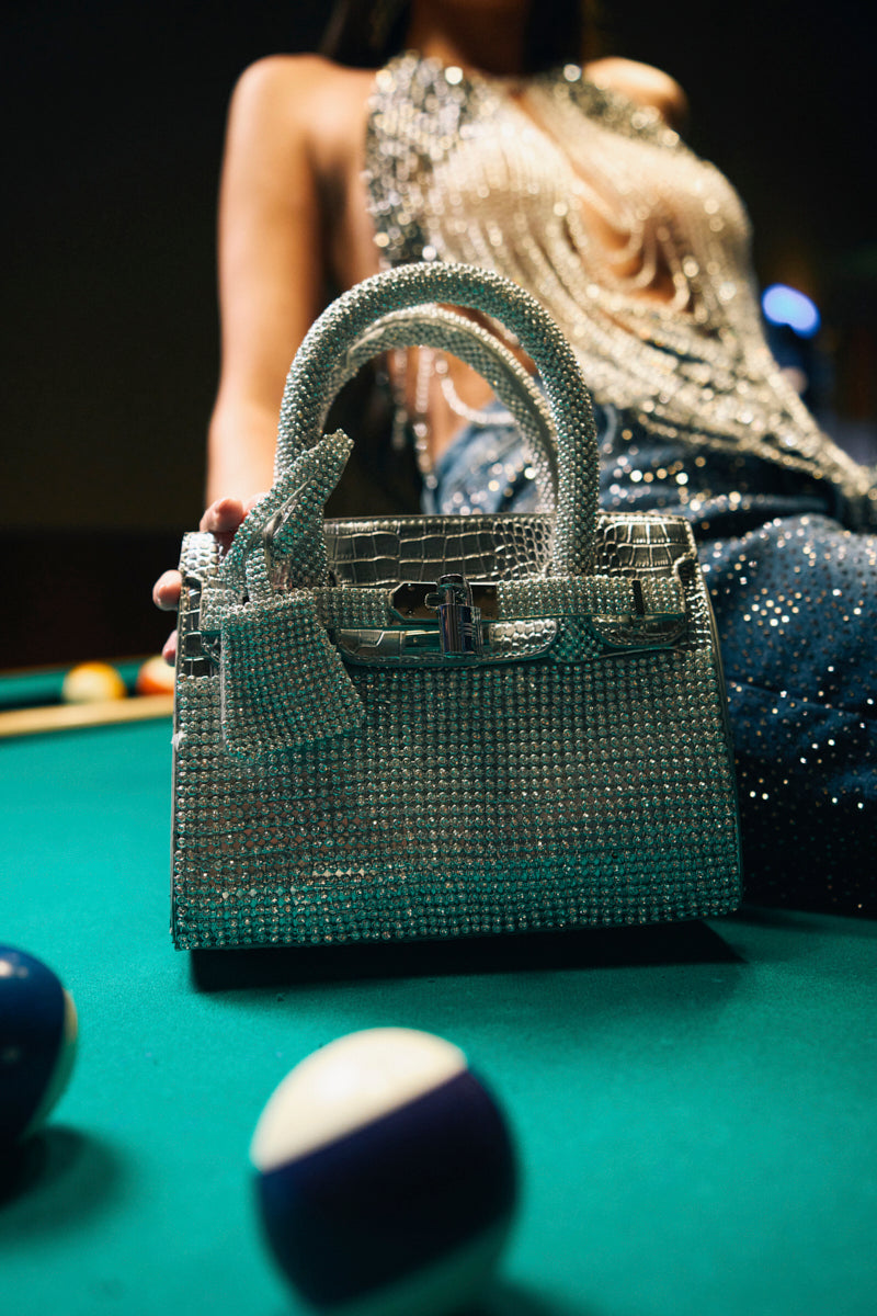 The SWERVE BLING PURSE, a silver rhinestone handbag with a faux leather finish, sparkles on a pool table next to billiard balls, while a person in a glittering outfit appears blurred in the background.