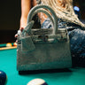 The SWERVE BLING PURSE, a silver rhinestone handbag with a faux leather finish, sparkles on a pool table next to billiard balls, while a person in a glittering outfit appears blurred in the background.