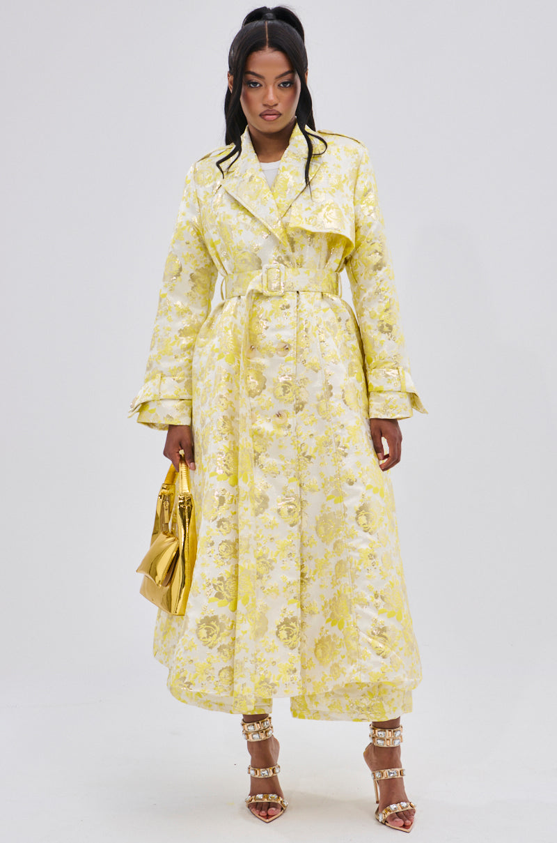 A woman poses against a plain background in the NO EXPECTATIONS BROCADE TRENCH—a long yellow coat with metallic floral motifs and double-breasted front—styled with gold high-heeled sandals and a shiny gold handbag.