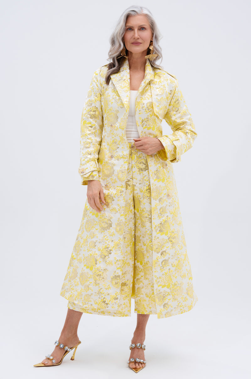 A woman with long gray hair wears the NO EXPECTATIONS BROCADE TRENCH with metallic floral patterns, double-breasted front, matching wide-leg trousers, and embellished heeled sandals, standing against a plain white background.