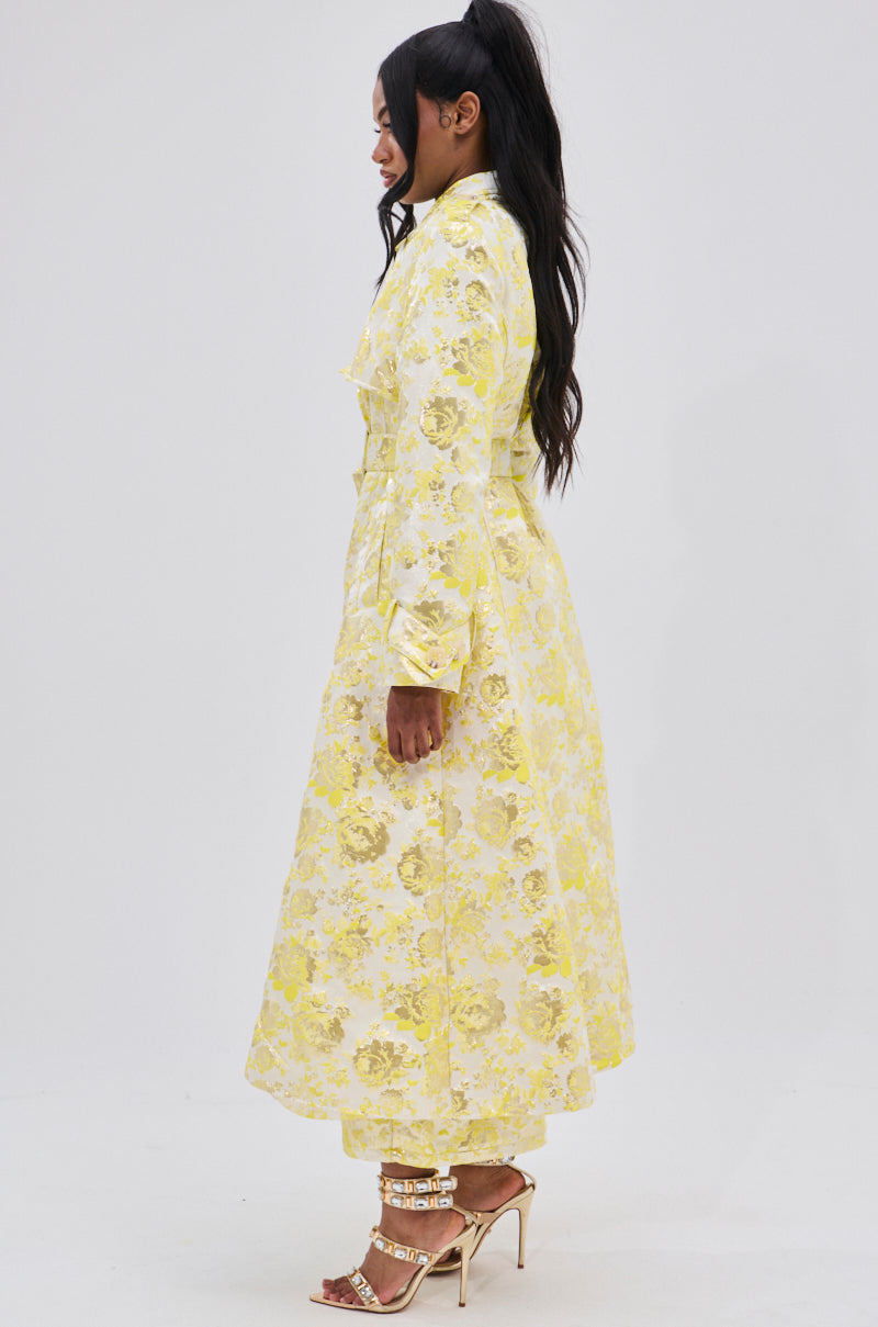 A woman in profile wears the NO EXPECTATIONS BROCADE TRENCH with metallic floral motifs over matching pants. She has long dark hair, strappy high heels, and stands against a plain white background.