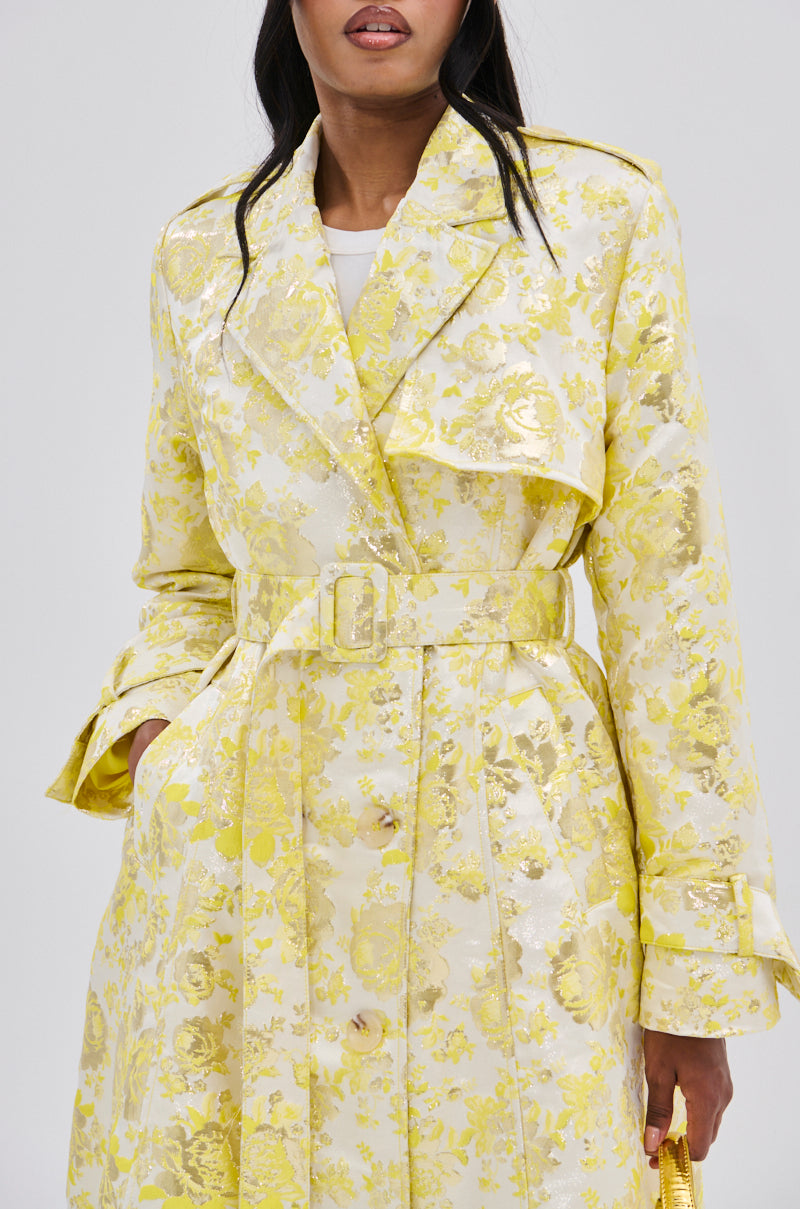 A woman wears the NO EXPECTATIONS BROCADE TRENCH, a light yellow coat with metallic floral motifs, belted waist, and large lapels. She holds a small yellow handbag against a plain background; her face is partly visible.
