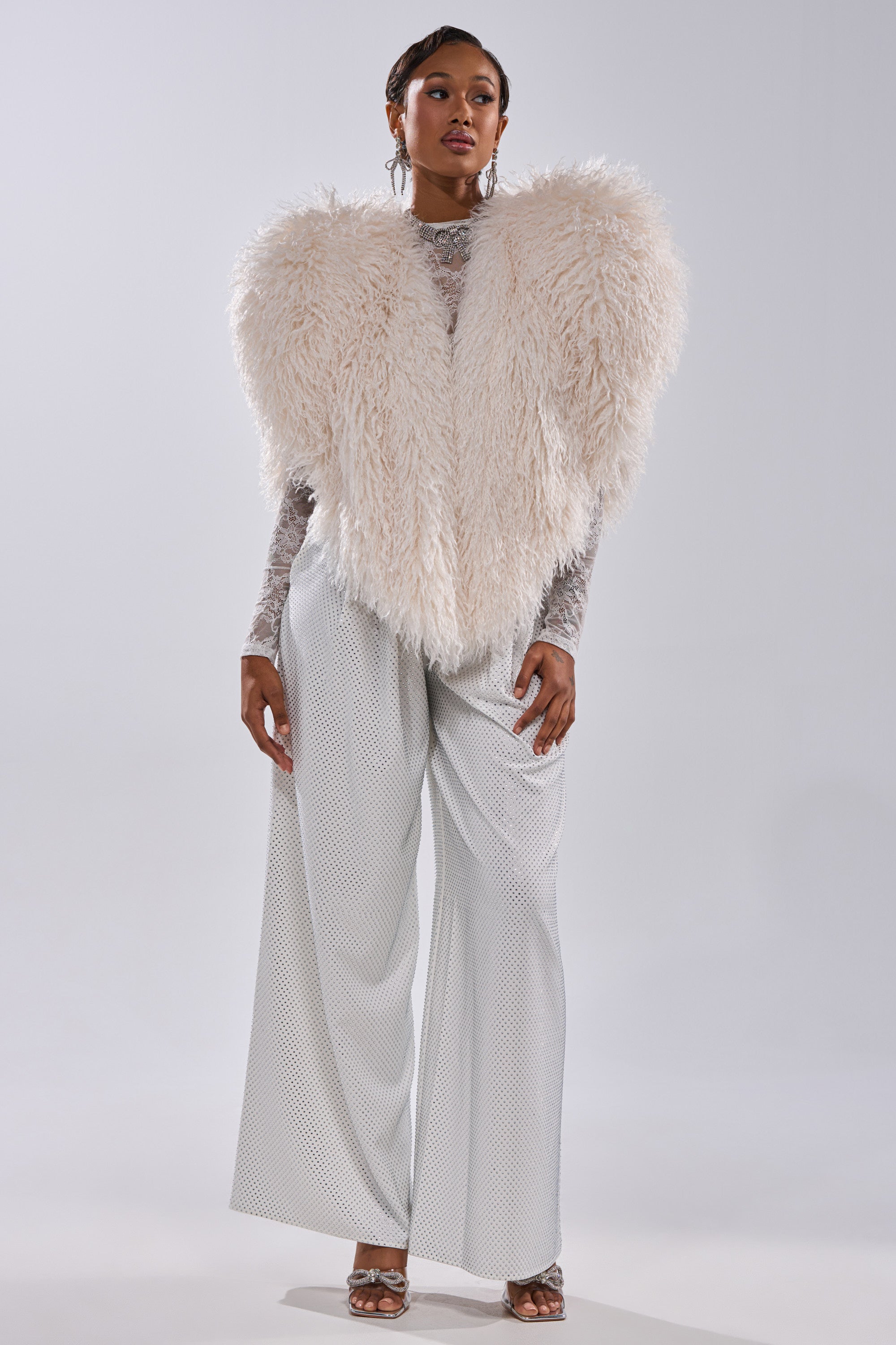 A woman poses confidently against a light gray background, wearing wide-legged silver pants, a sheer silver top, and the STAR CROSSED LOVER FAUX FUR VEST IN IVORY as a statement piece.