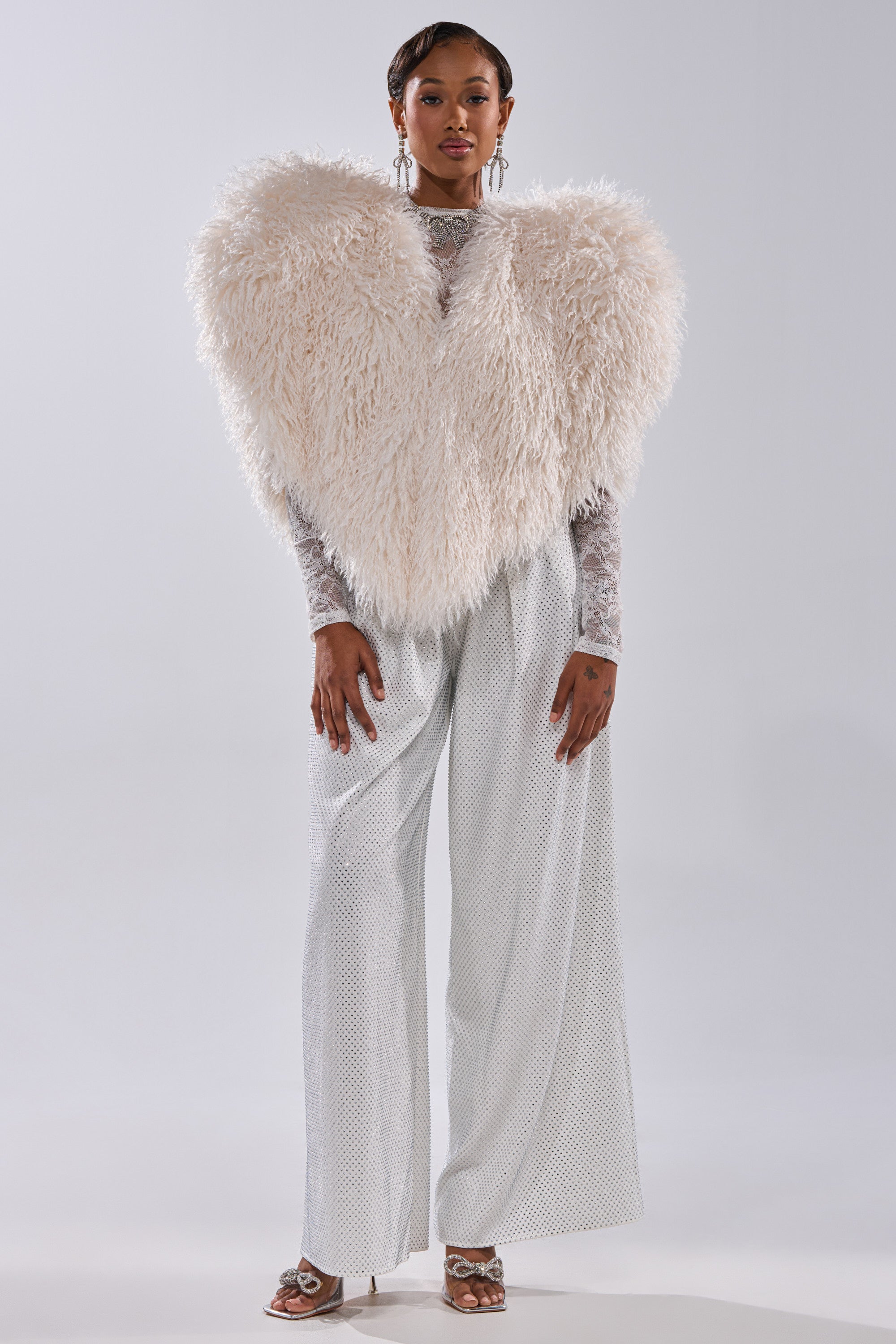 A woman models the STAR CROSSED LOVER FAUX FUR VEST IN IVORY as a statement layer over a sheer long-sleeve top and wide-legged, shimmery silver pants with matching silver open-toe heels, posed against a plain light backdrop.