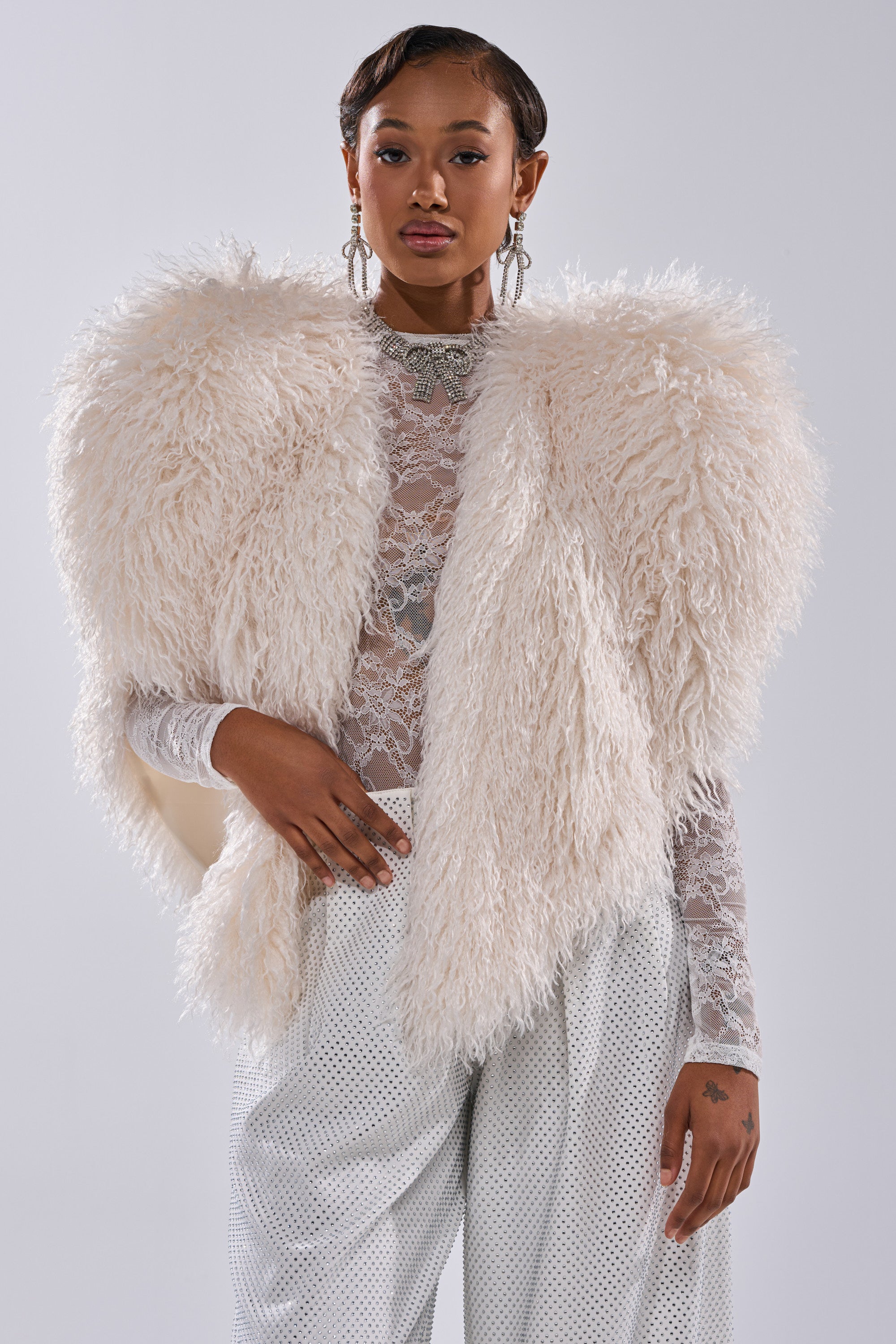 A confident person in the STAR CROSSED LOVER FAUX FUR VEST IN IVORY layers it over a sheer lace top and loose white pants, accessorizing with bold jewelry to make the vest stand out against a plain backdrop.
