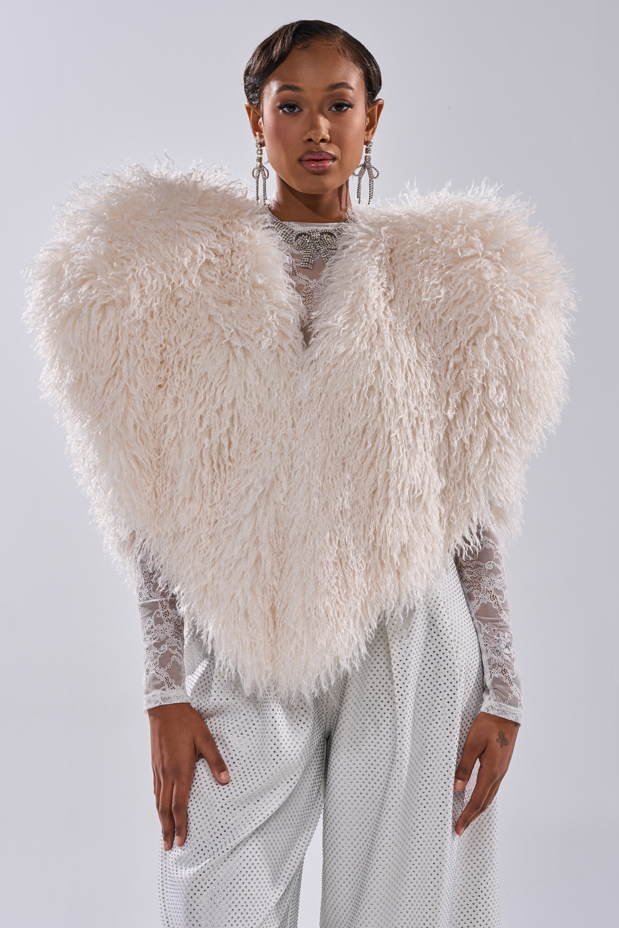 A woman with short, styled hair and statement earrings wears the STAR CROSSED LOVER FAUX FUR VEST IN IVORY over a lace top and high-waisted, wide-leg pants, standing against a plain background.