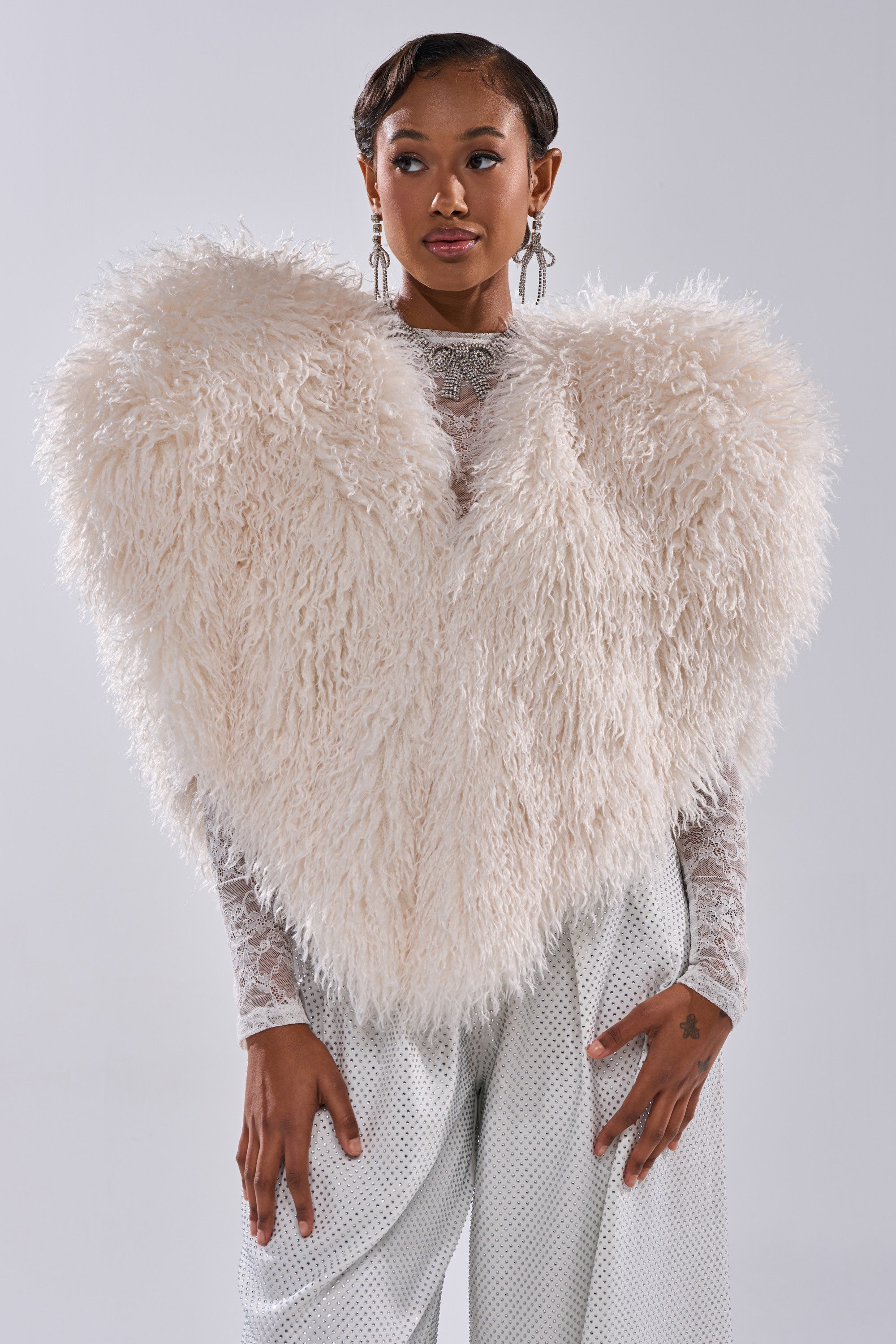 A woman models the STAR CROSSED LOVER FAUX FUR VEST IN IVORY—a bold, fluffy statement—over a sheer lace top and white pants, accessorized with chic earrings and a sleek hairstyle, set against a simple background.