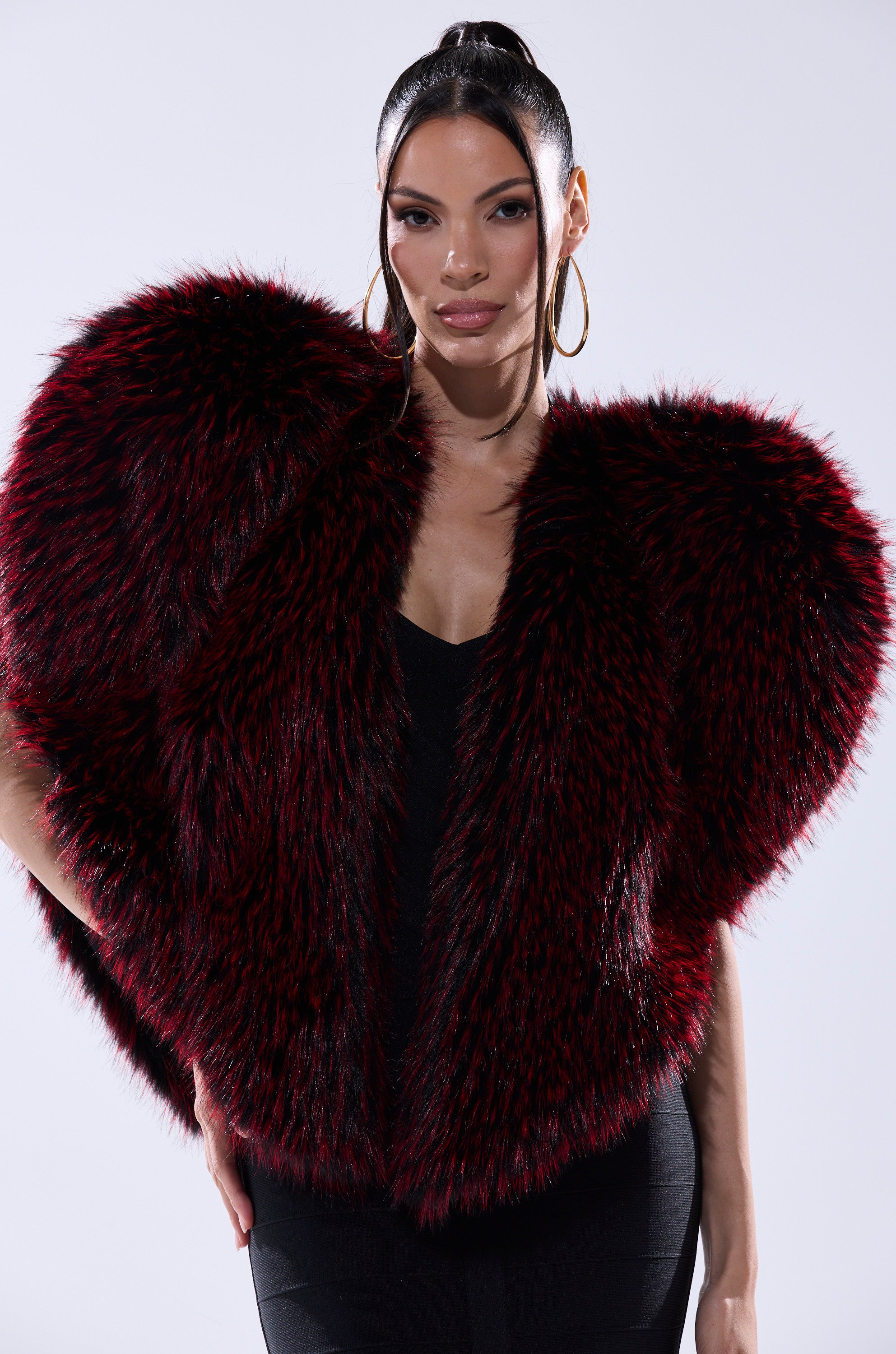 A woman with long dark hair tied back wears large gold hoop earrings, a black dress, and the bold STAR CROSSED LOVER FAUX FUR VEST shaped like a heart. She poses confidently against a light gray background, making it a statement piece.