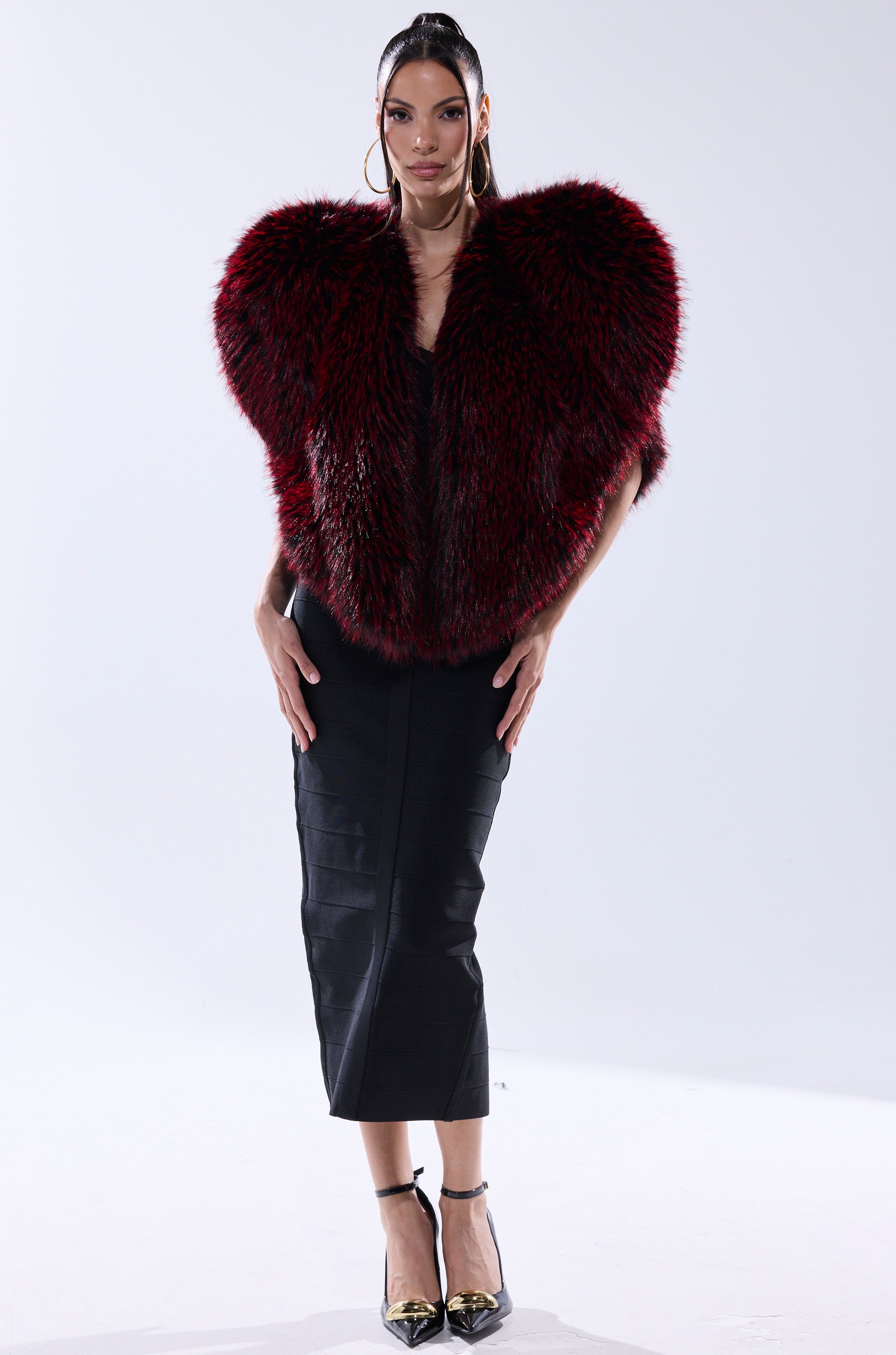 A woman poses against a white background in a black dress and gold heels, styled with the STAR CROSSED LOVER FAUX FUR VEST in deep red and black. She wears hoop earrings, has long hair, and exudes confidence.