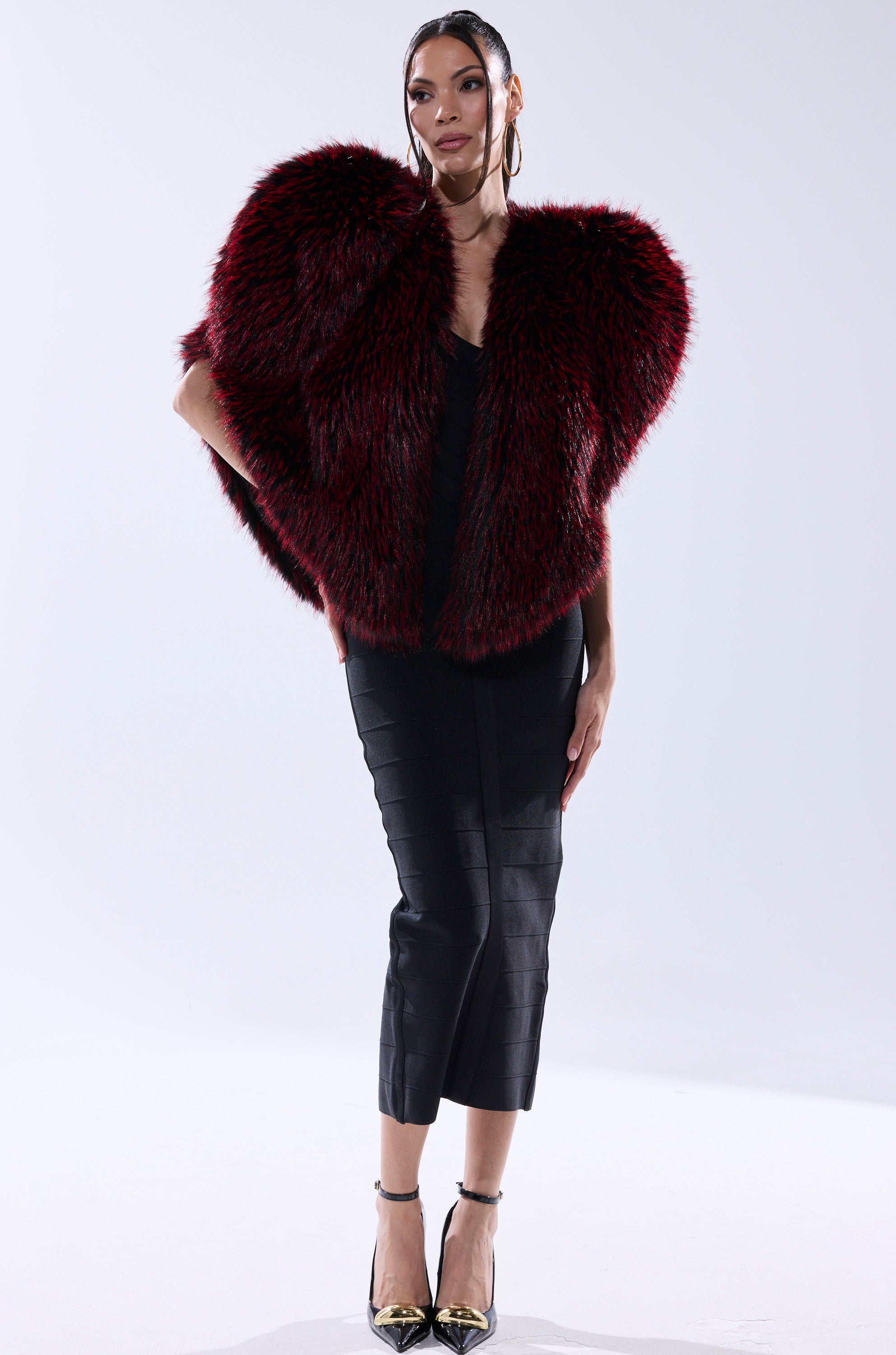 A woman poses in a black midi dress and metallic pointed heels, layering the STAR CROSSED LOVER FAUX FUR VEST—a deep red and black faux fur piece with a dramatic silhouette—against a plain white background.