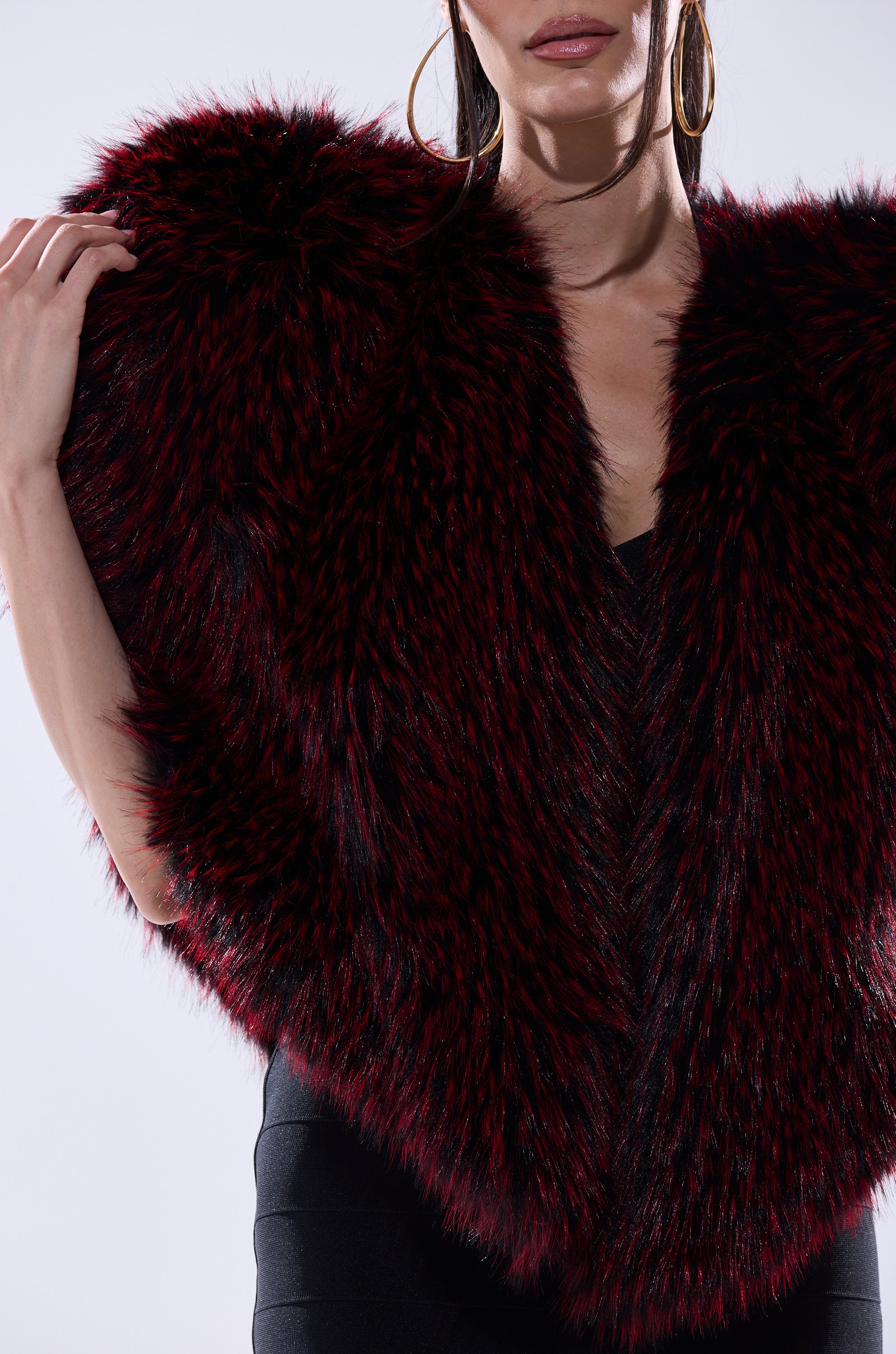 A woman models the STAR CROSSED LOVER FAUX FUR VEST in deep red and black over a black sleeveless dress with large hoop earrings, shown from shoulders to mid-thigh against a plain background.