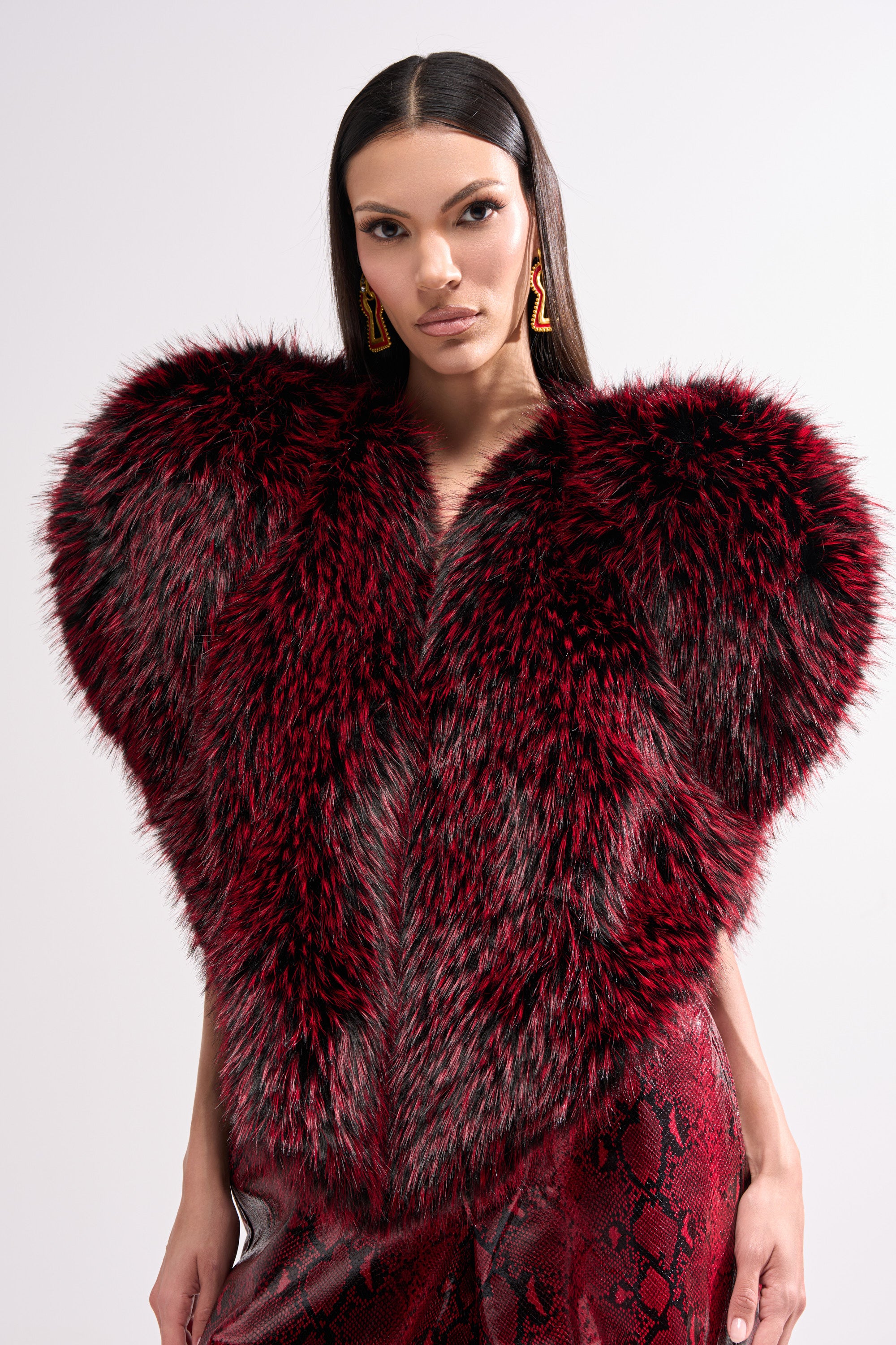 A woman with straight dark hair wears the dramatic STAR CROSSED LOVER FAUX FUR VEST in red and black, bold zigzag earrings, and red snakeskin-patterned pants, standing against a plain light background.