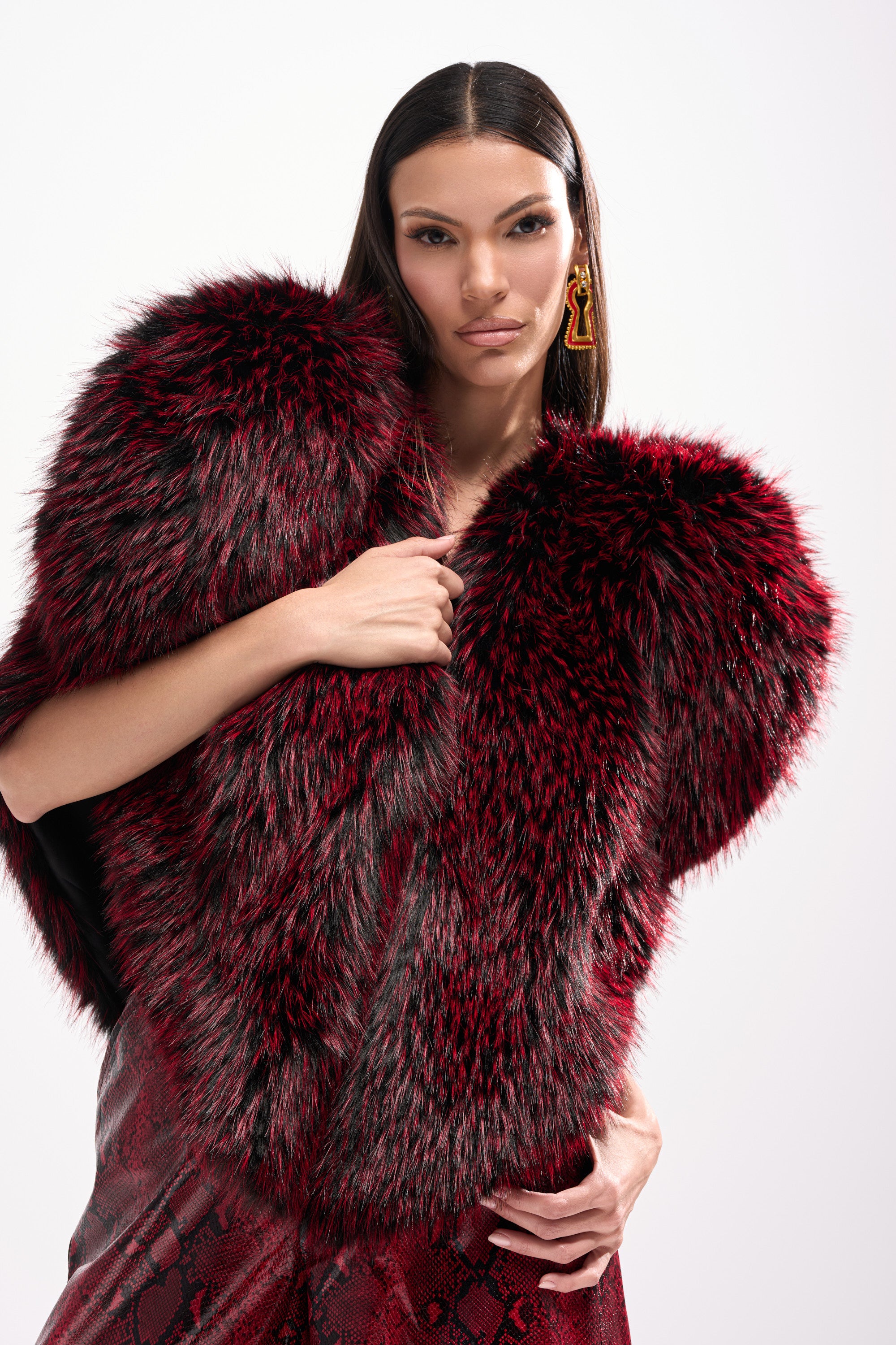 A woman with long dark hair poses confidently in the STAR CROSSED LOVER FAUX FUR VEST, a luxurious red and black faux fur piece, paired with red patterned pants against a plain white background.